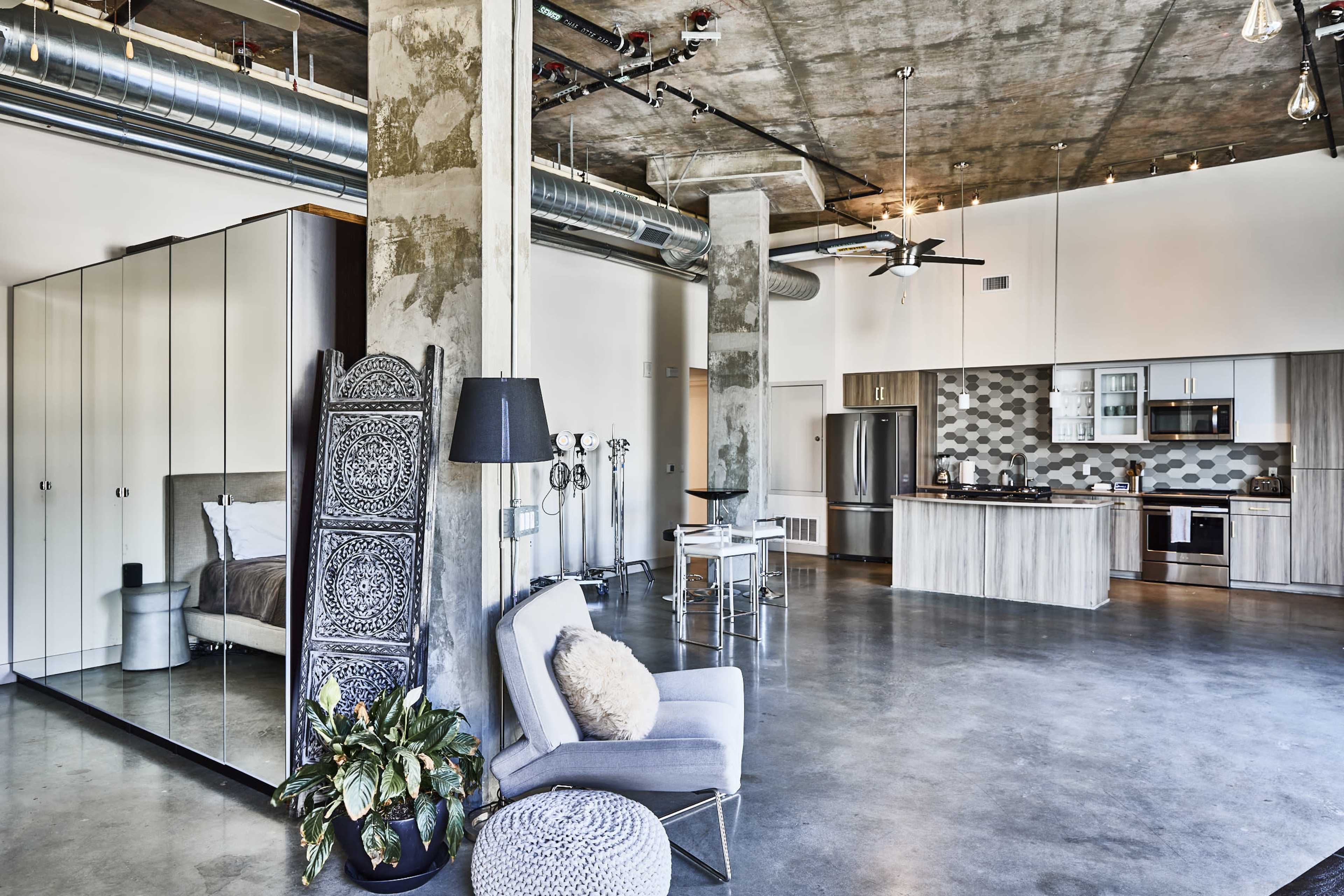The image shows a modern, industrial-style apartment featuring an open living space with a kitchen, seating area, and large windows.