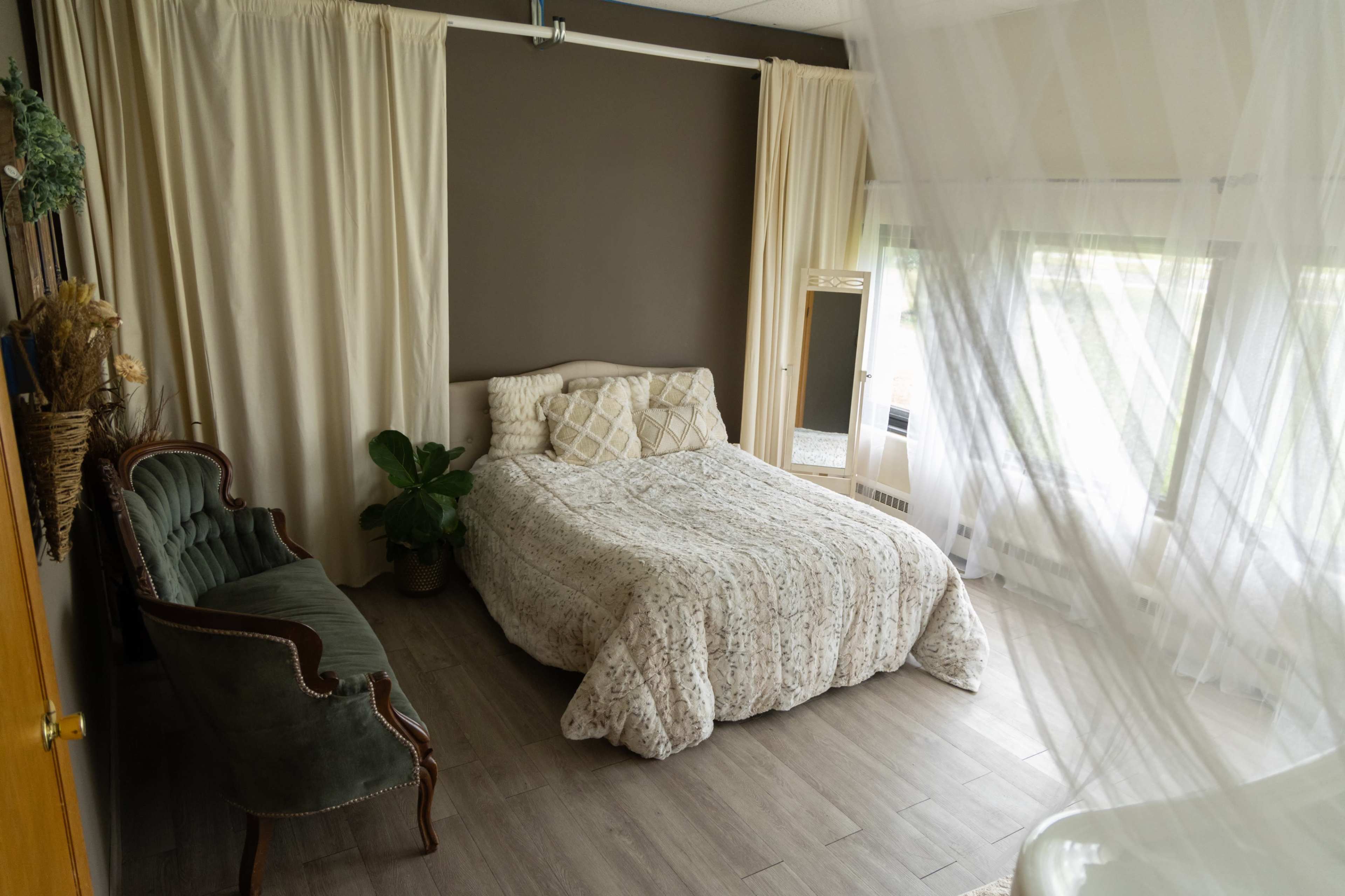 The image shows a simple bedroom with a bed covered in a patterned blanket, a decorative chair, and natural light filtering through sheer curtains.