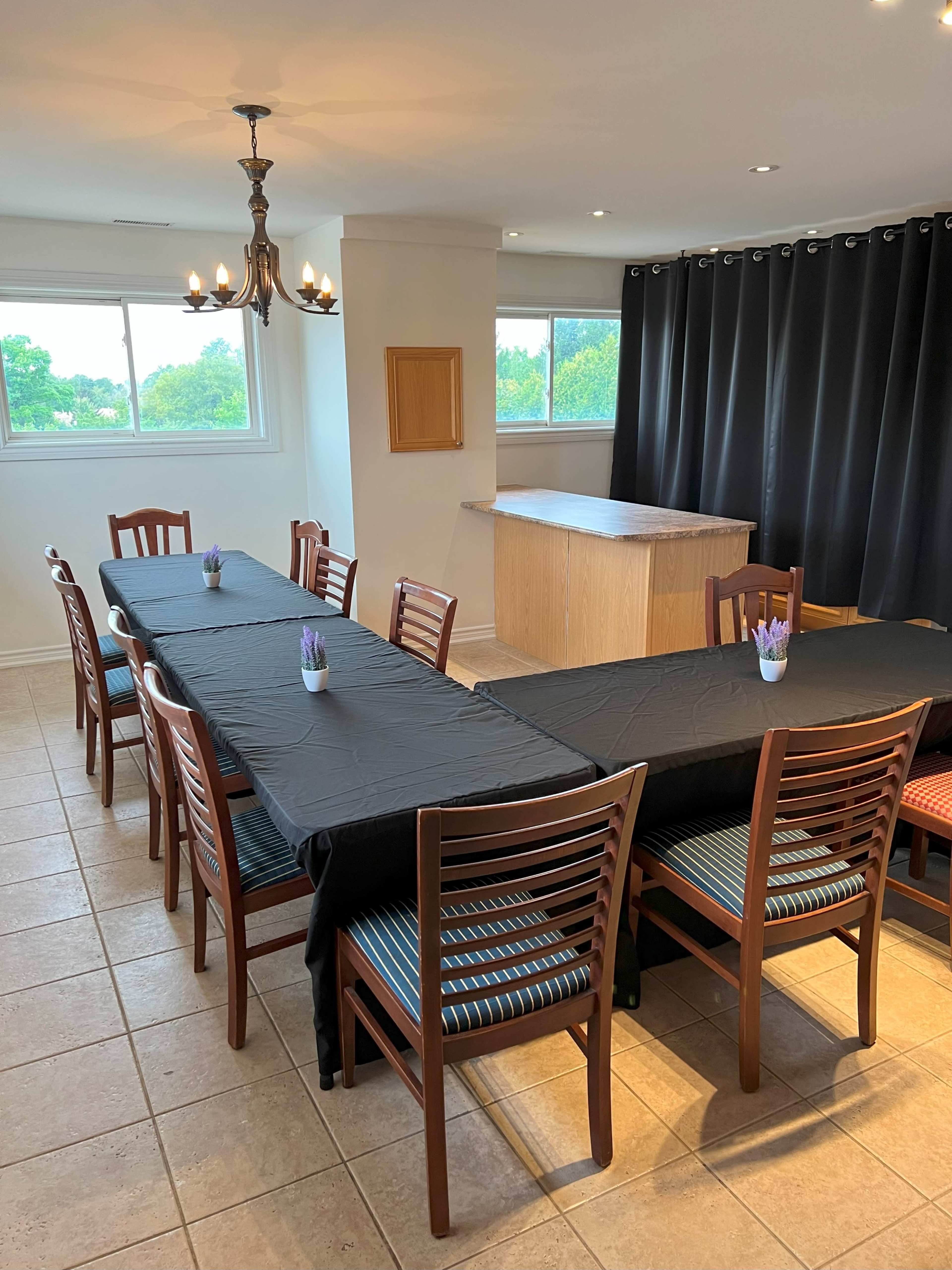 A long dining table with black tablecloths and wooden chairs is set in a well-lit room with large windows and dark curtains.