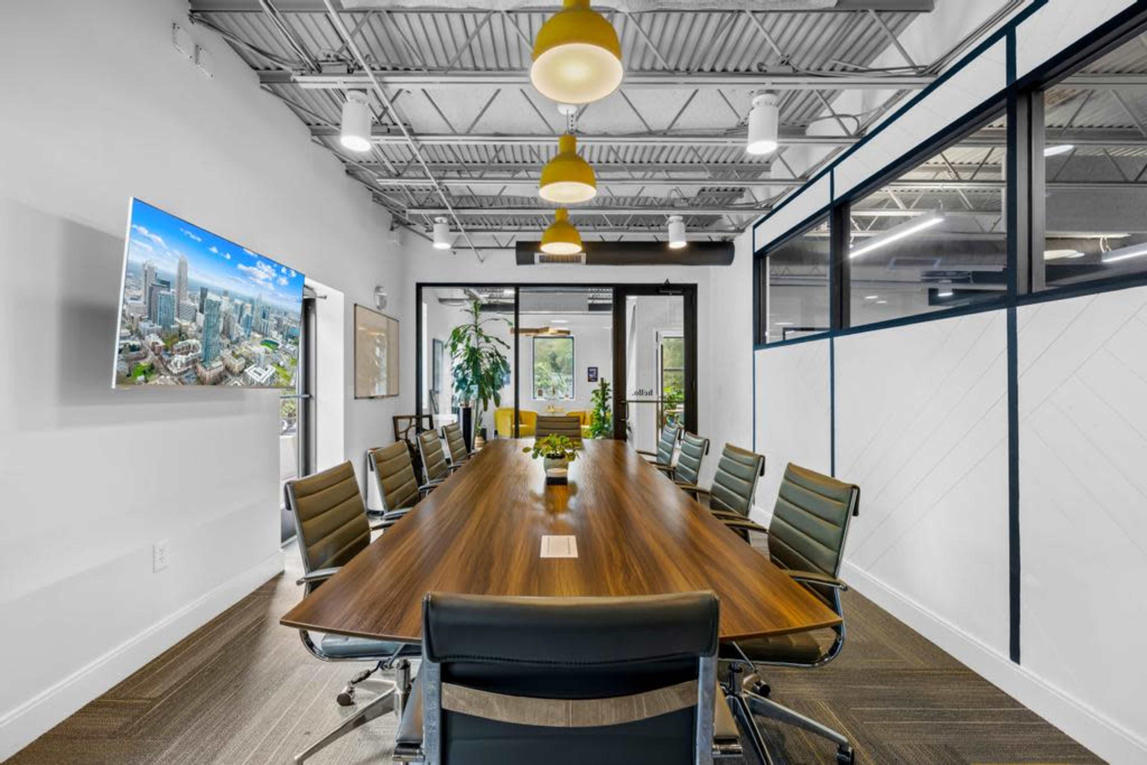 A modern conference room features a long wooden table surrounded by black leather chairs, with a large screen displaying a cityscape and natural light coming through windows.