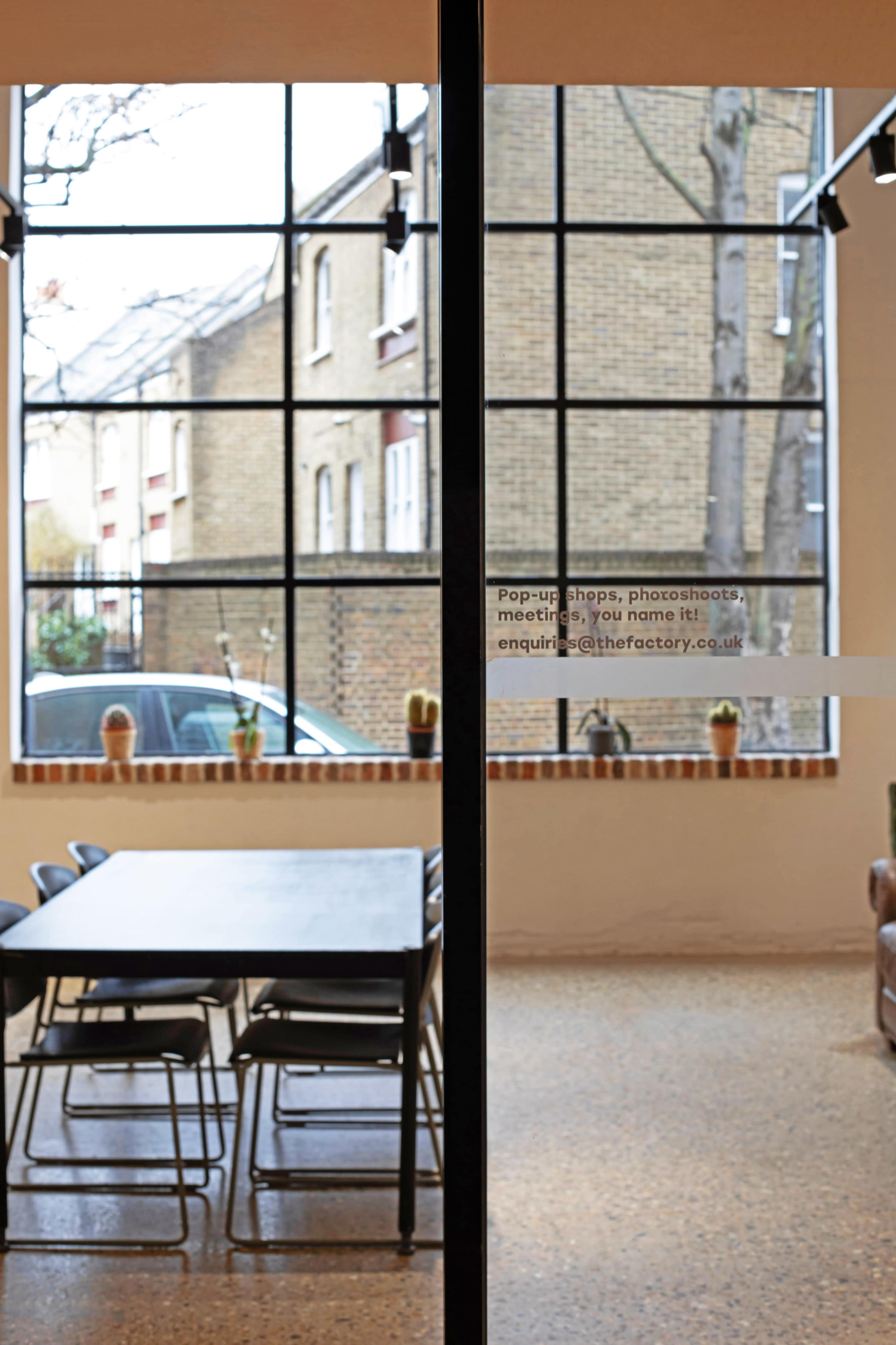 A table and chairs are set up in a room with large windows overlooking a brick building and outdoor plants.