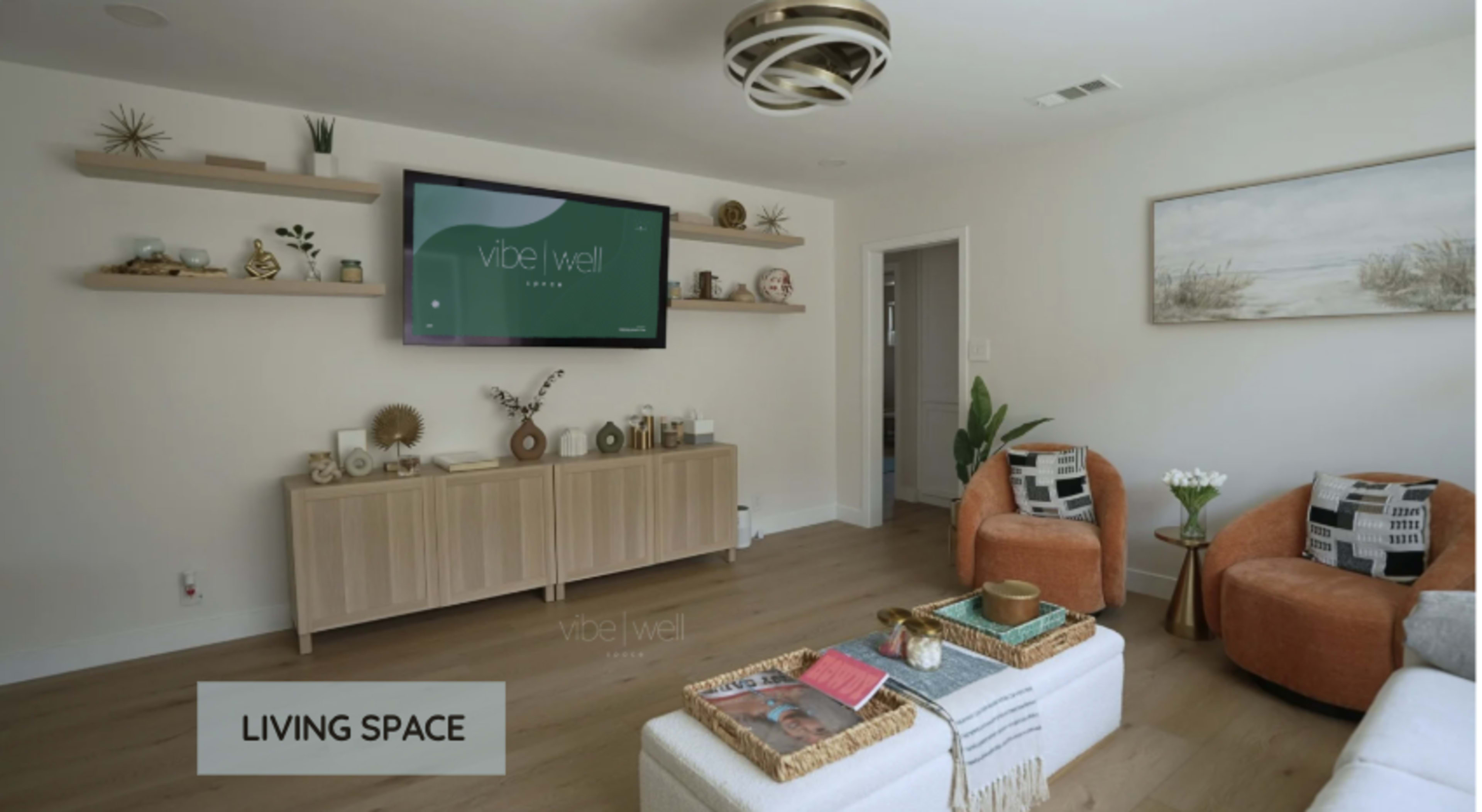 A living room features a mounted television, wooden shelves displaying decorative items, and two orange accent chairs facing a coffee table with magazines.