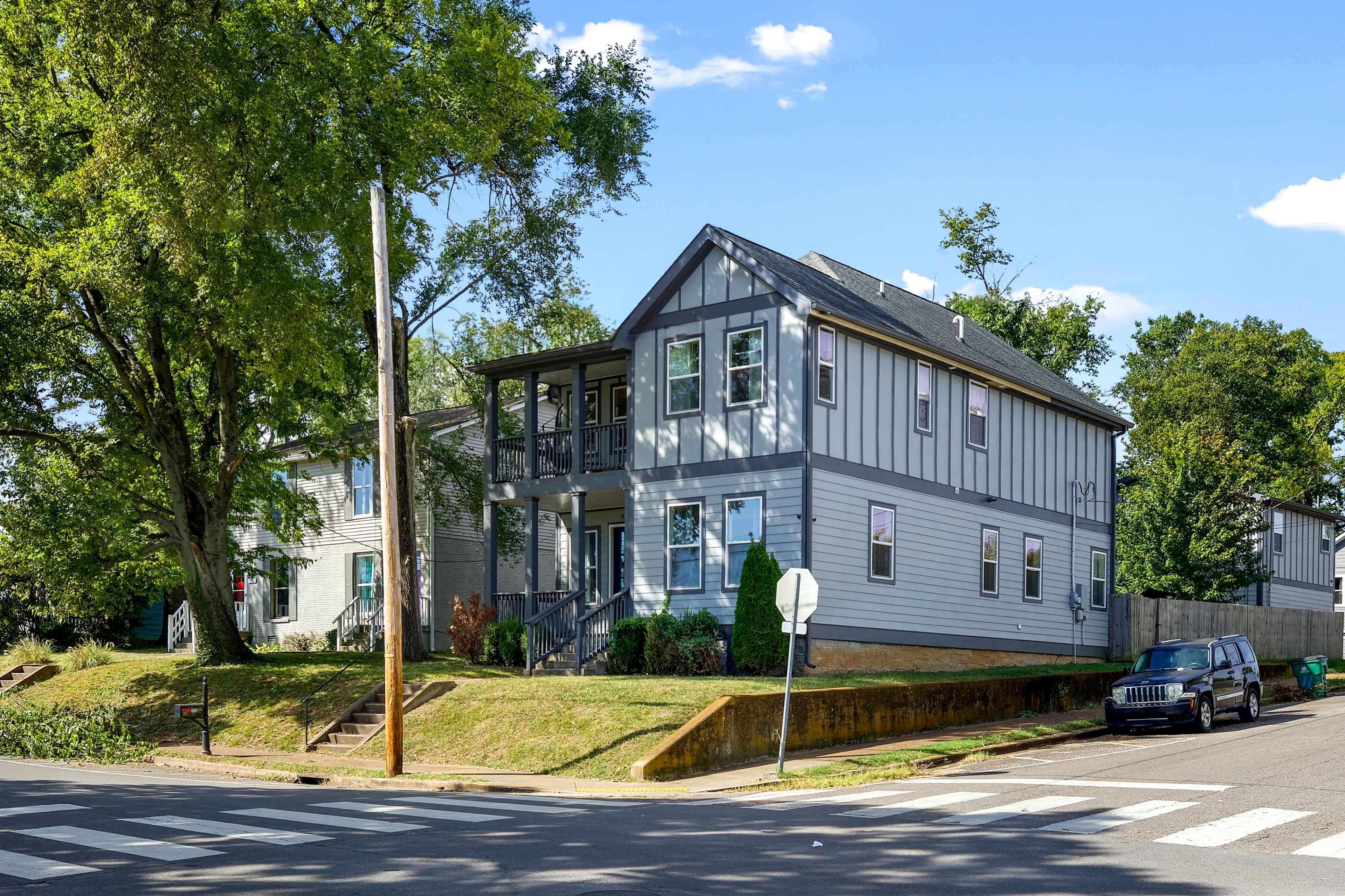 A two-story gray house with a porch, situated at the corner of a street with parked cars and greenery nearby.