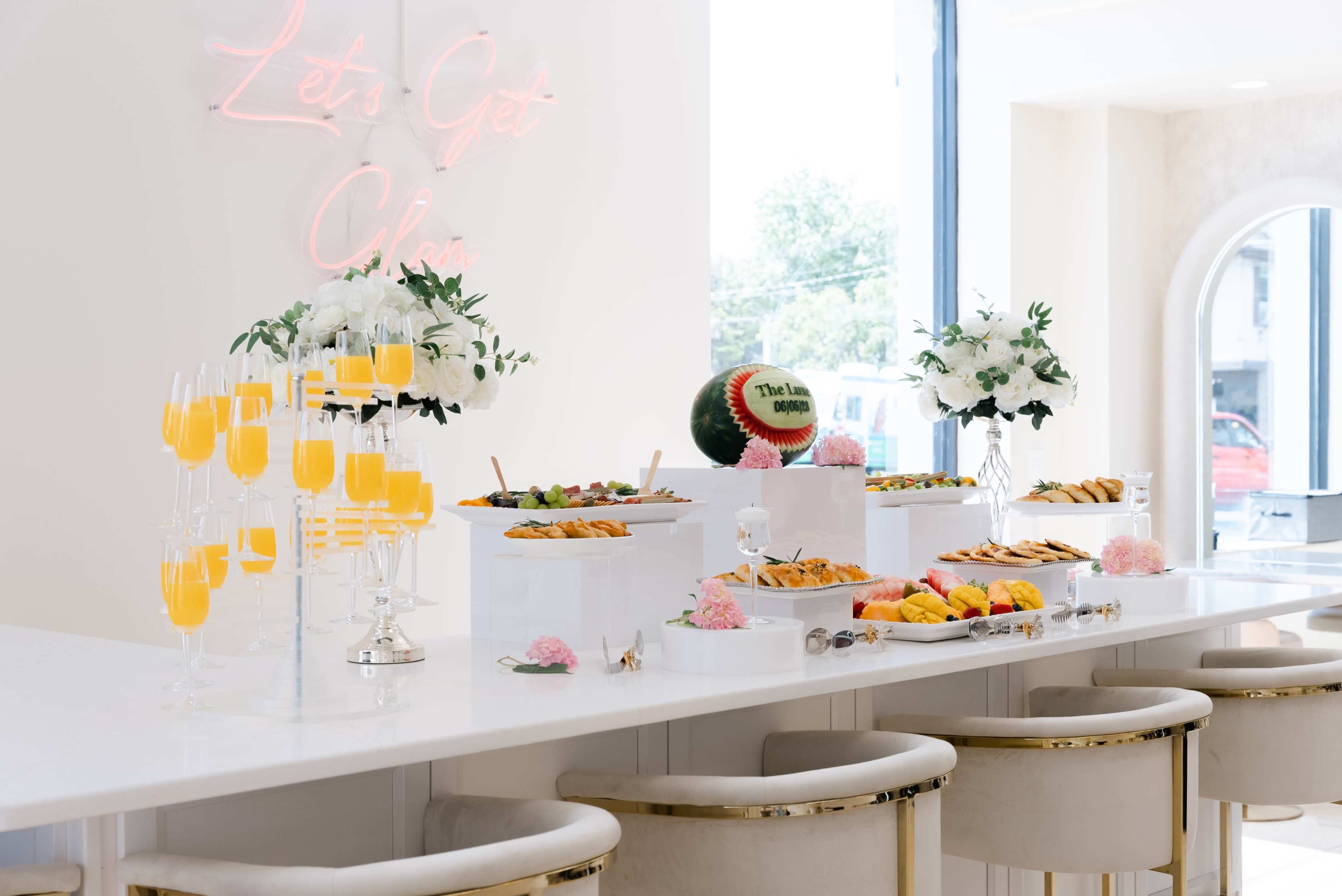 A bright, modern event space features a buffet table filled with various appetizers and drinks, accented by a neon sign and floral arrangements.