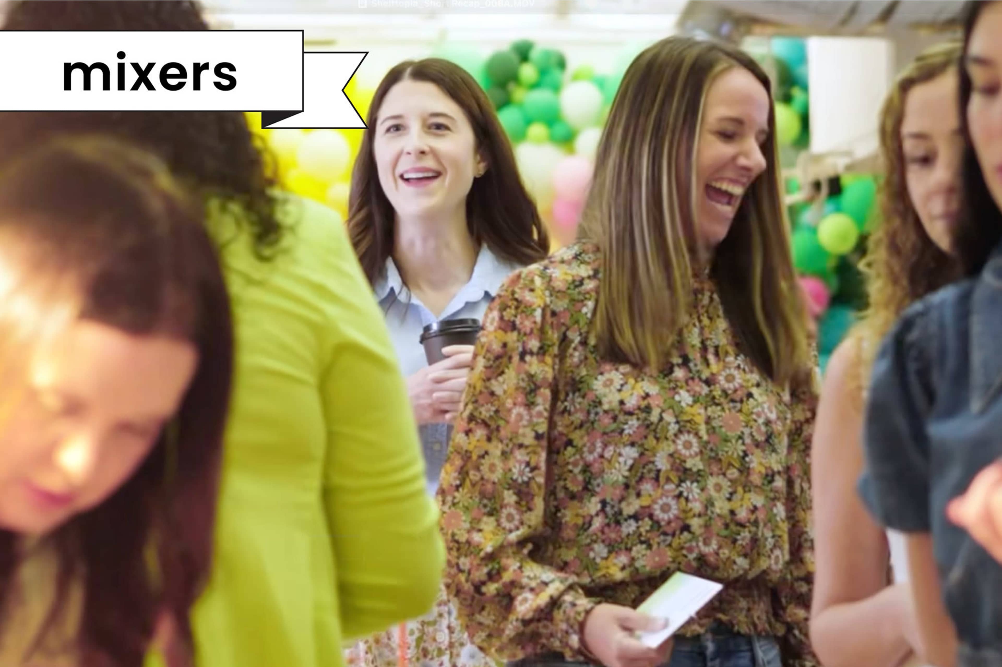 A group of women are socializing at an event surrounded by colorful decorations and chatting while one woman holds a coffee cup.