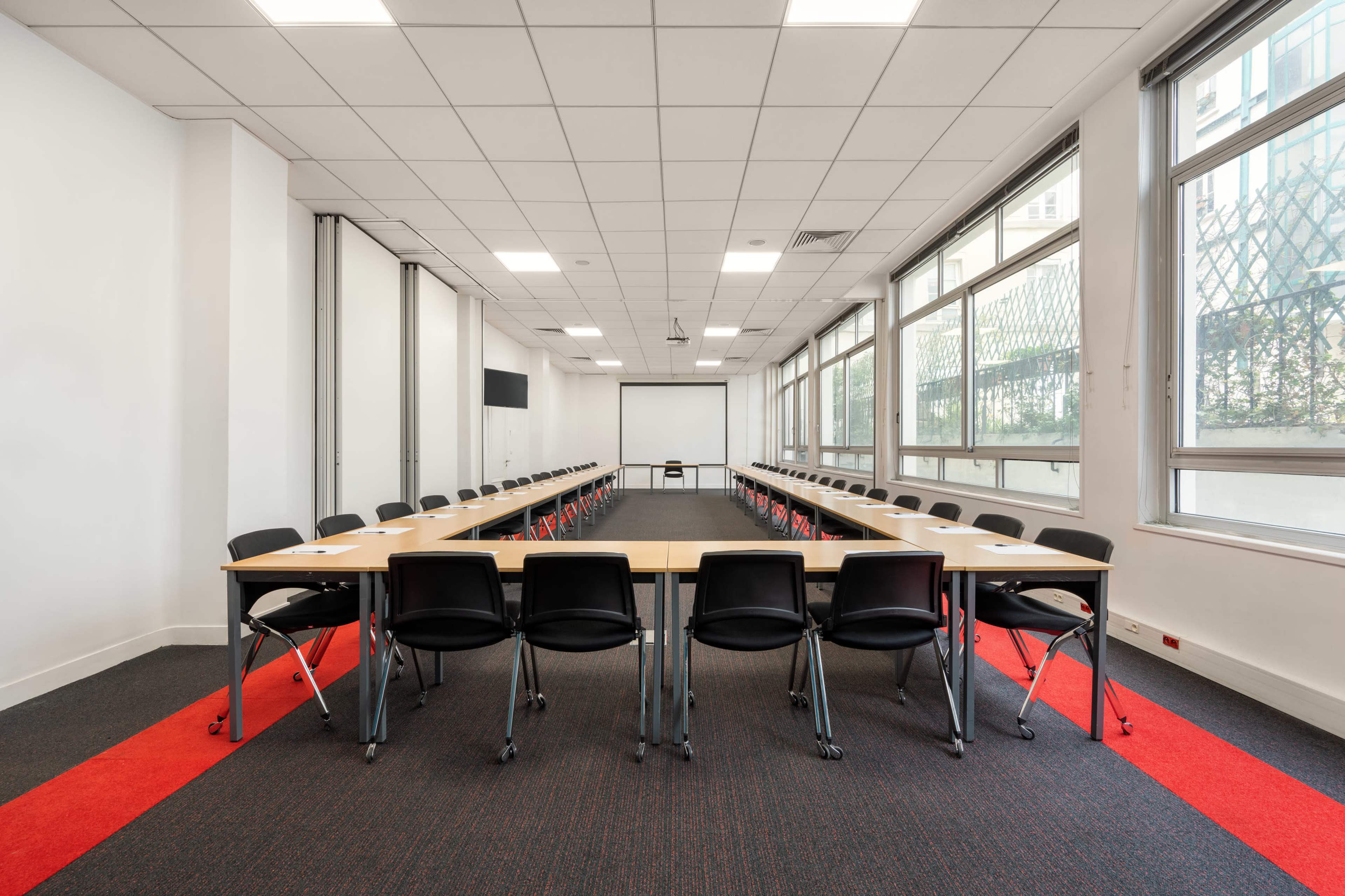 A rectangular conference room is arranged with tables and chairs set in a U-shape, featuring large windows and a red carpet in the center.