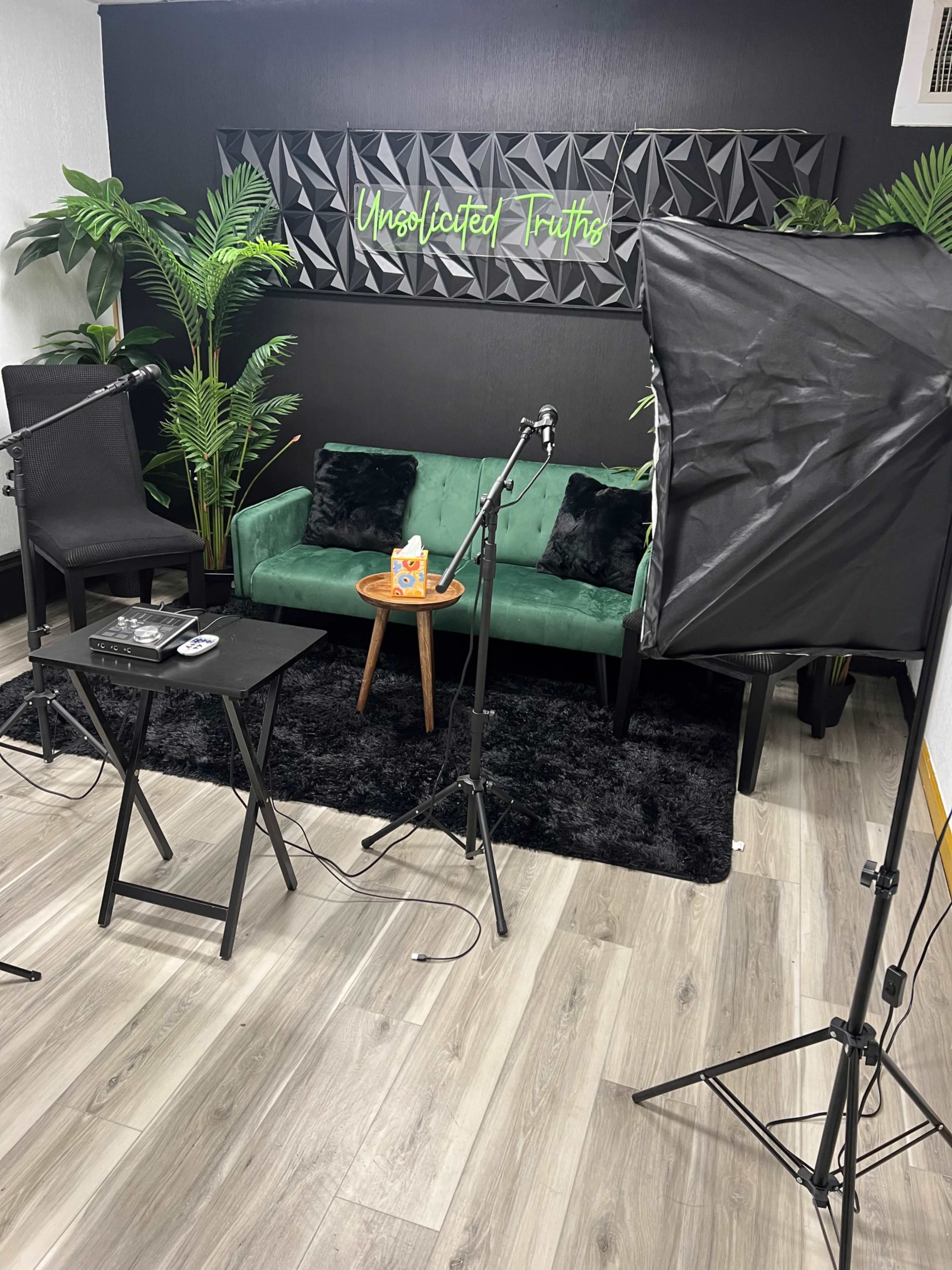A podcast or interview setup featuring a green couch, a small table with a snack, two microphones, and studio lighting against a black wall with the words "Unsolicited Truths."