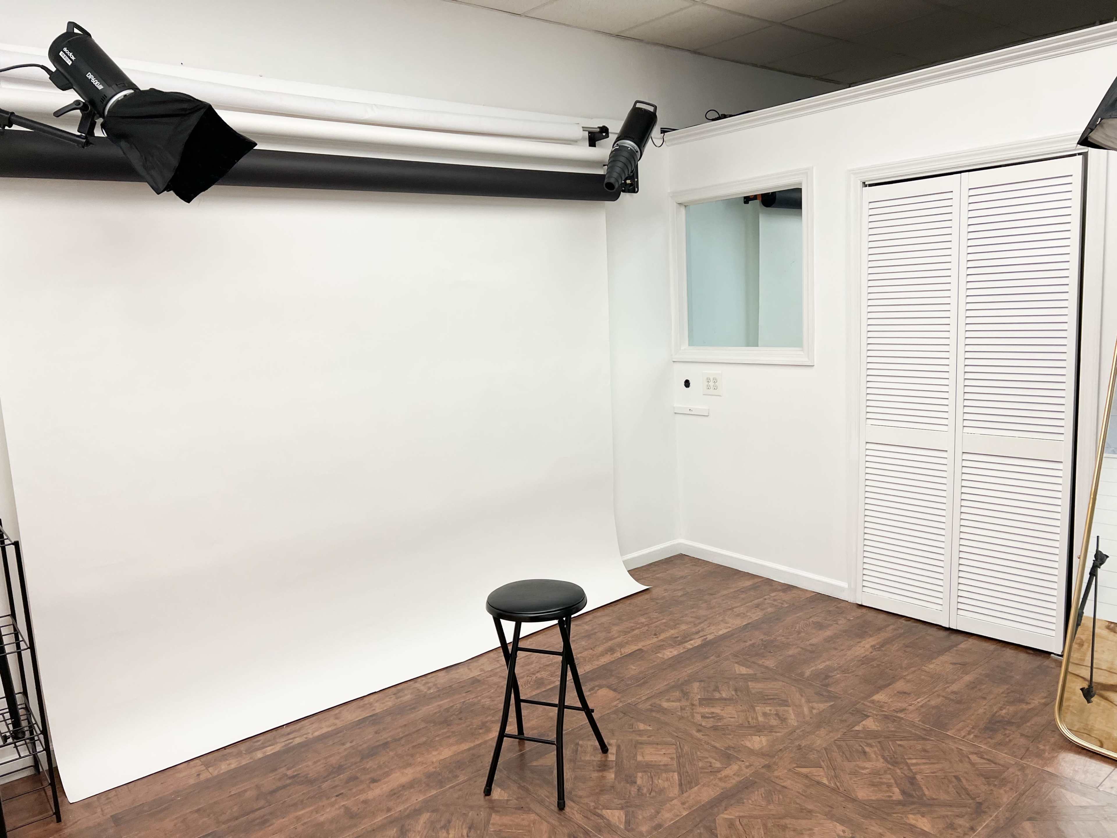 A photography studio with a plain white backdrop, a black stool, and various lighting equipment mounted overhead.