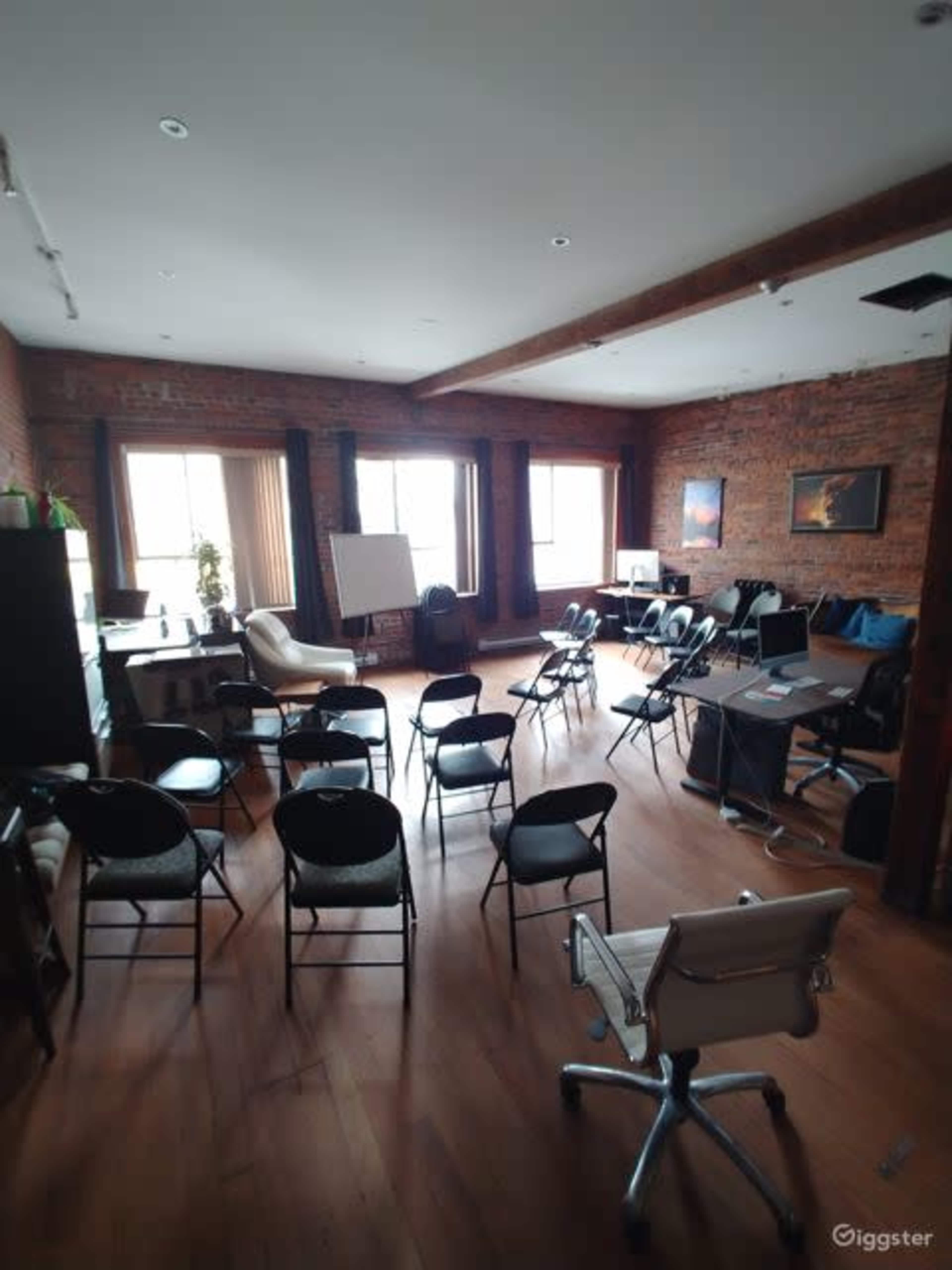 The image shows a spacious meeting room with wooden floors, brick walls, and various seating arrangements including folding chairs and a desk setup.