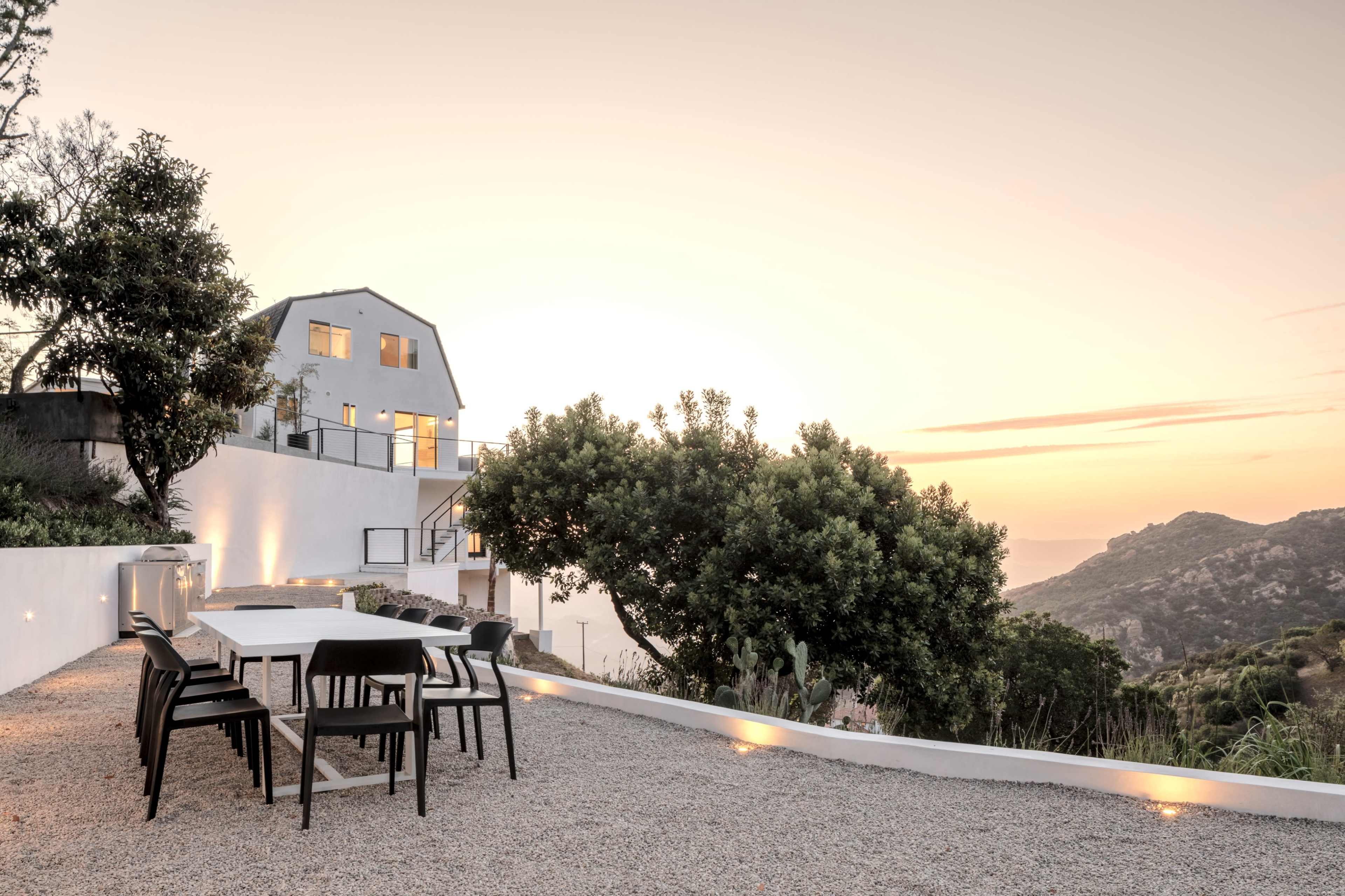 A modern house with large windows overlooks a gravel patio equipped with a long dining table and chairs, set against a sunset backdrop.