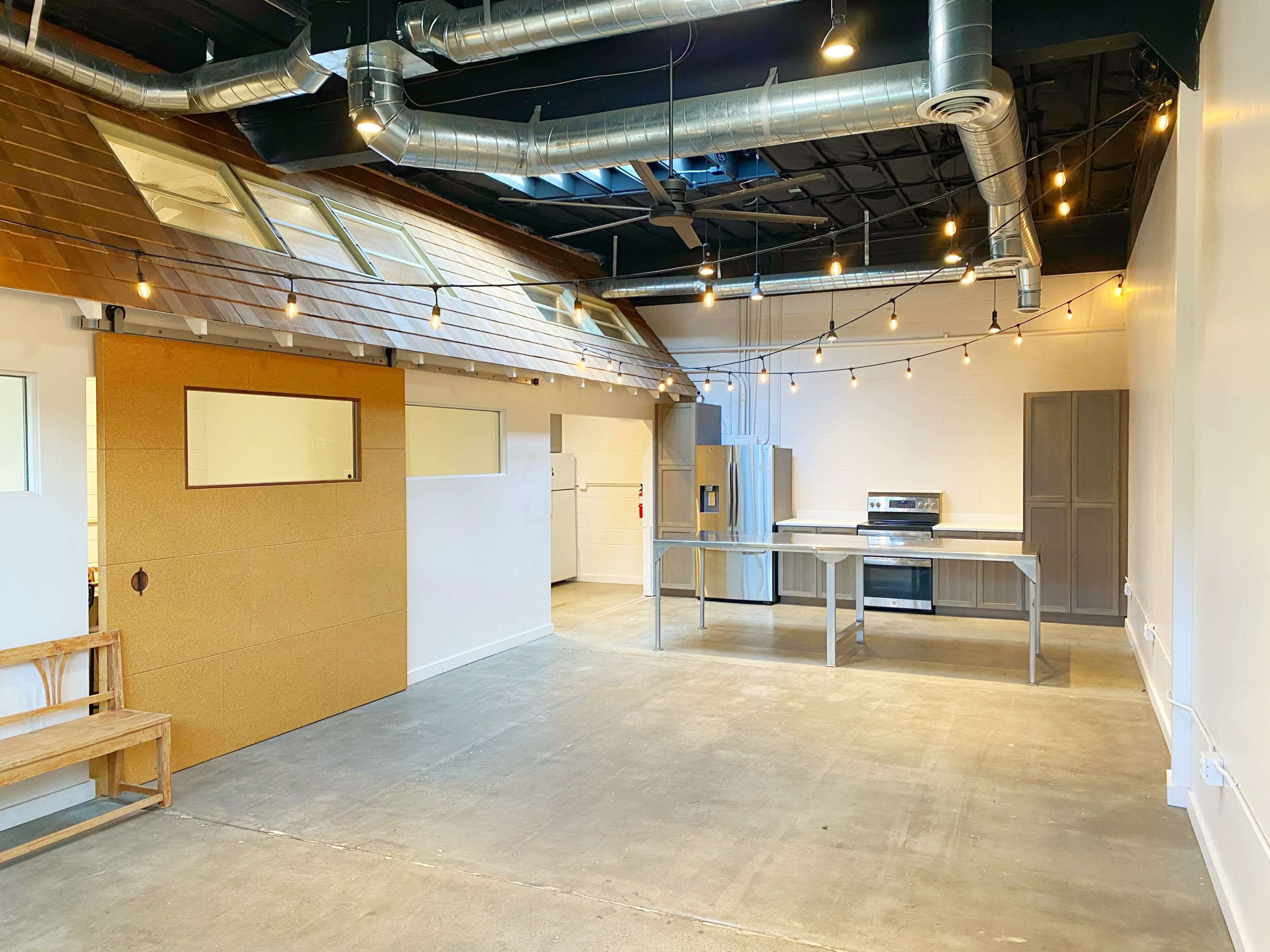 A spacious, modern kitchen area with stainless steel appliances, a wooden bench, and overhead string lights in an industrial-style room.