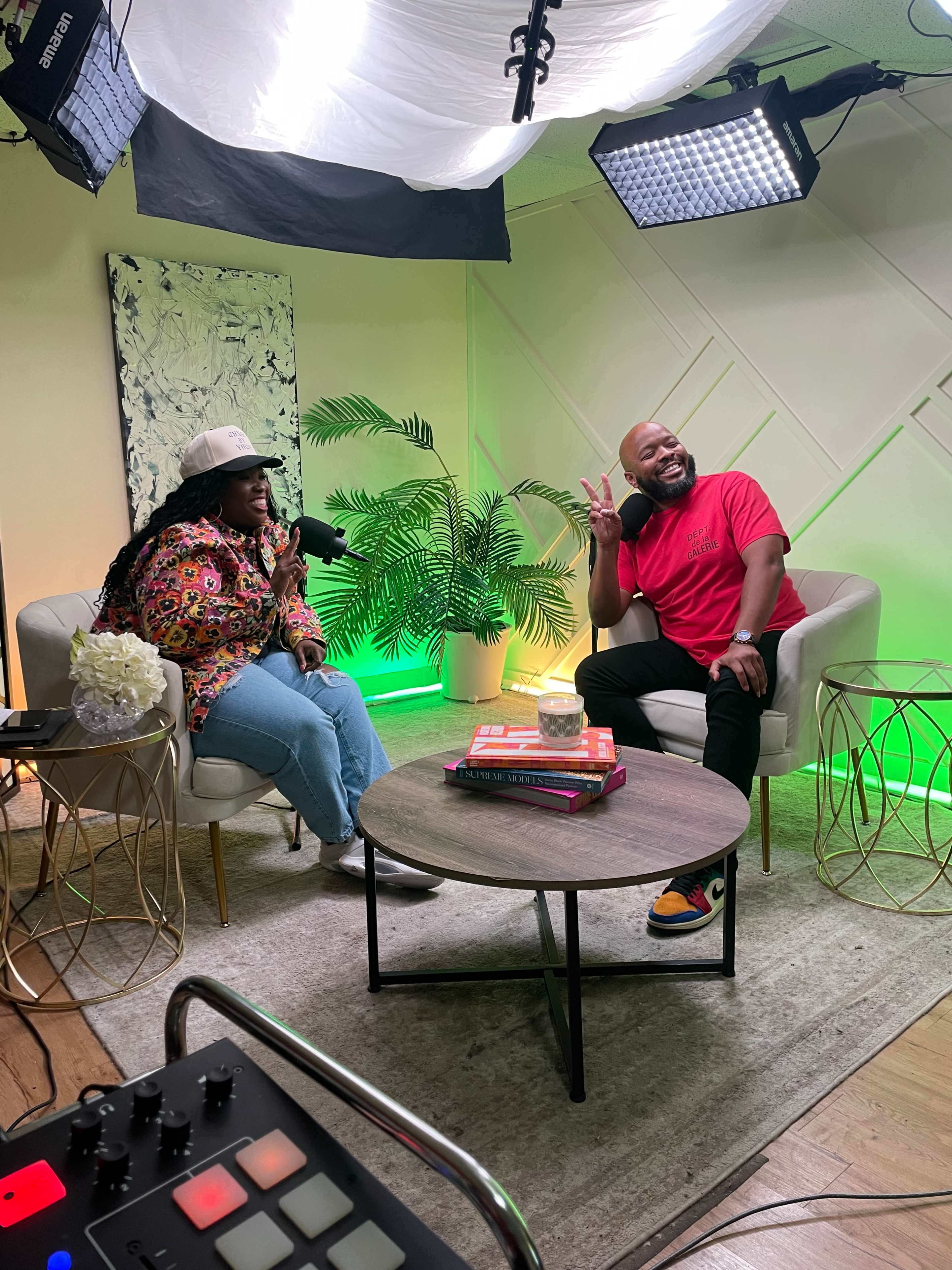 A woman and a man are seated in a modern studio setup, discussing into microphones, with a tropical plant and colorful lighting in the background.