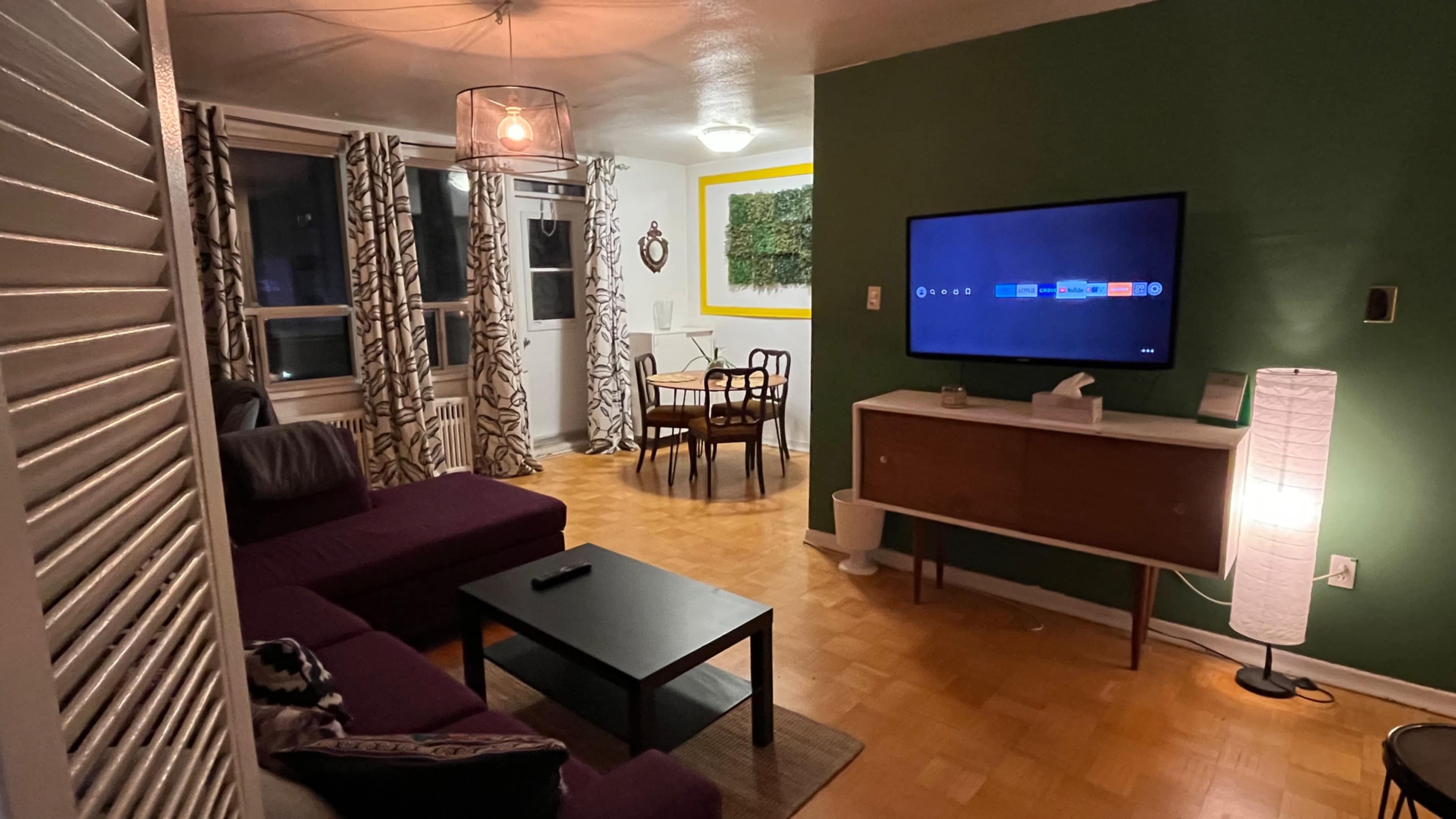 A living room with a purple couch and black coffee table, adjacent to a dining area with a round table and chairs, set against a green wall and light wood flooring.