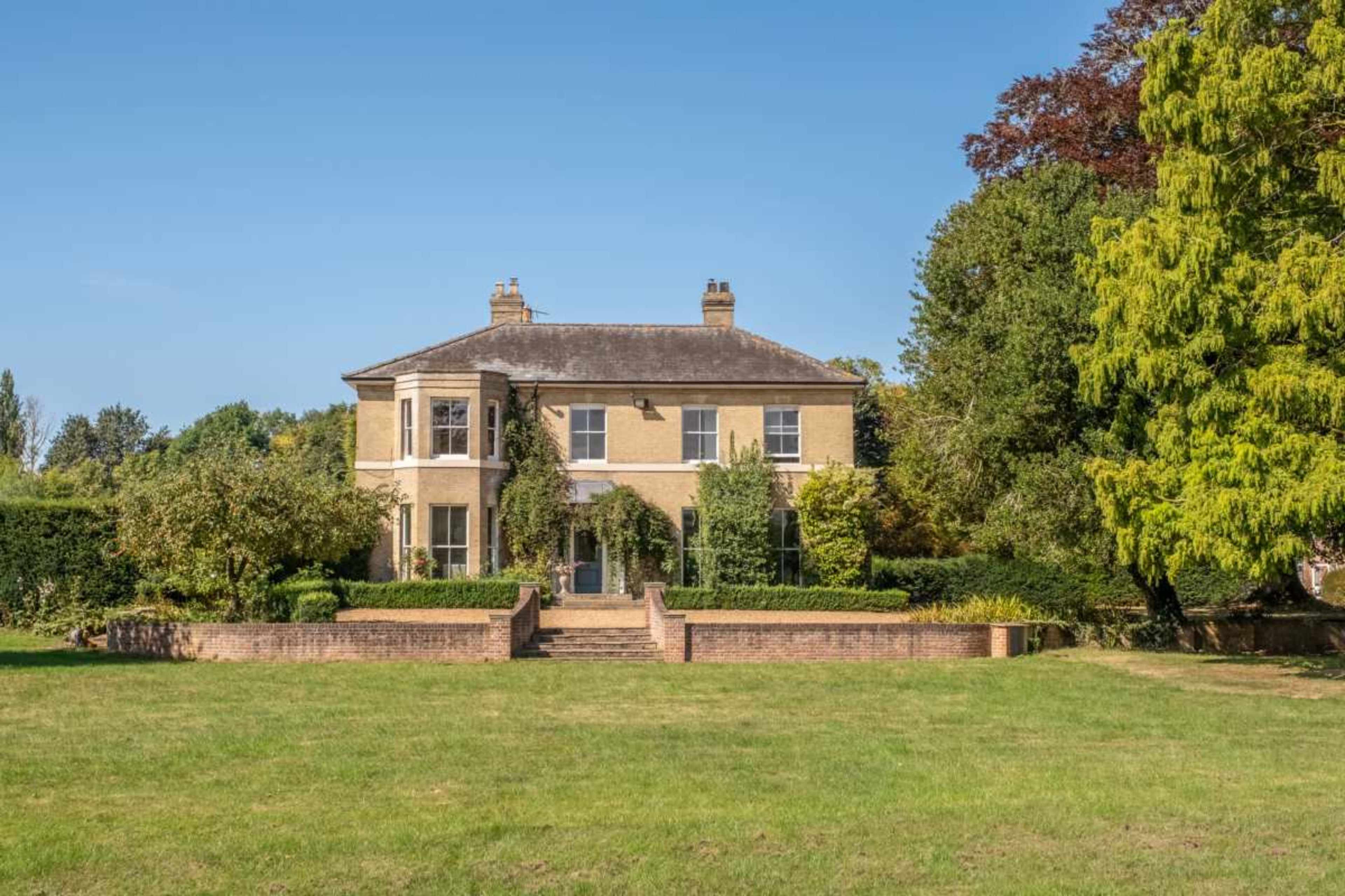 A large, two-story brick house is set back on a green lawn, surrounded by trees and low hedges.