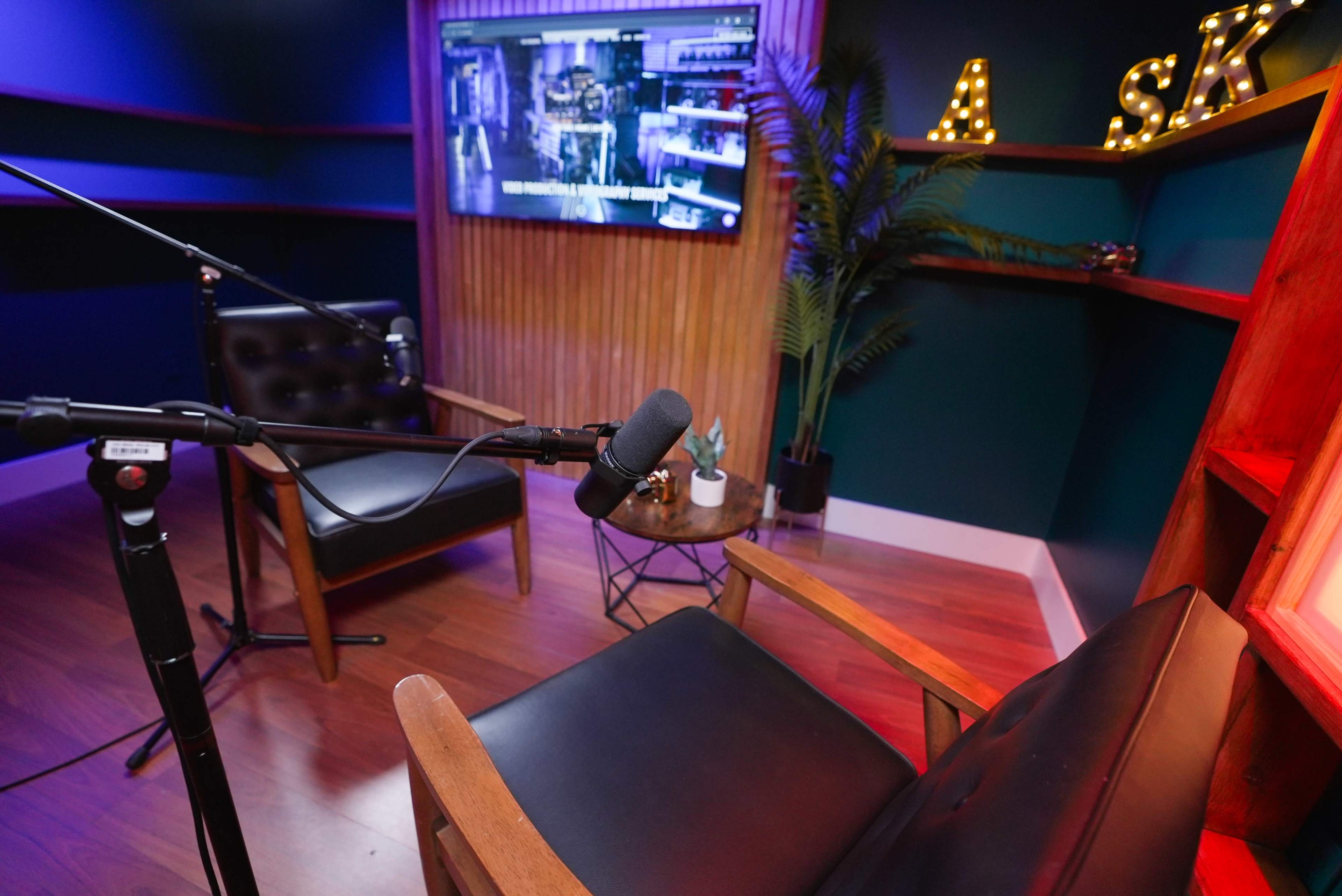 The image shows a small studio setup with two chairs, a microphone on a stand, and a television displaying a scene in the background.