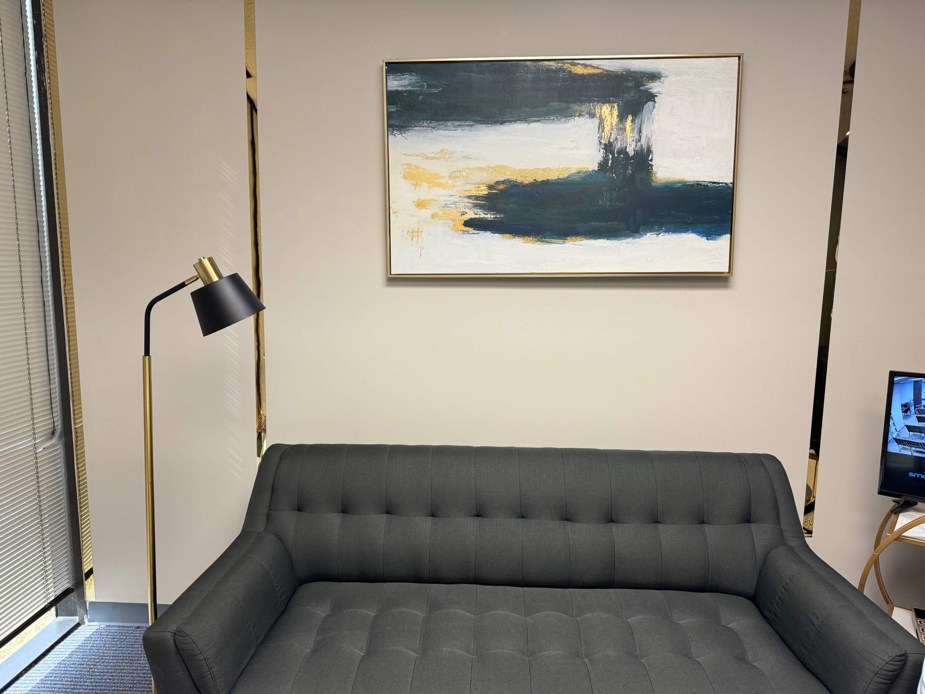 The image shows a modern office interior featuring a gray tufted sofa, a floor lamp, and an abstract painting hung on the wall.