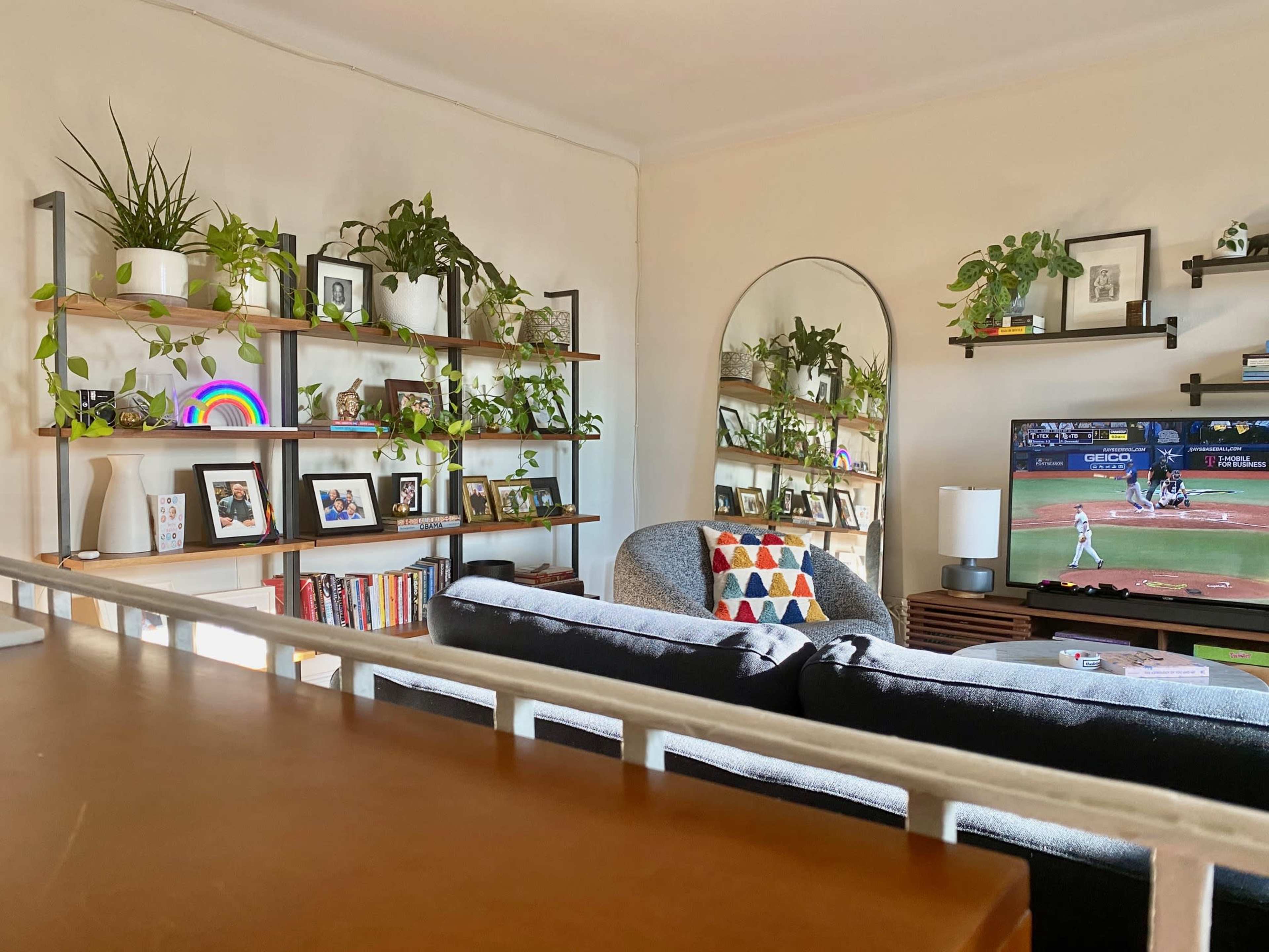 A living room features a cozy seating area, shelves filled with plants and photos, and a television displaying a sports event.