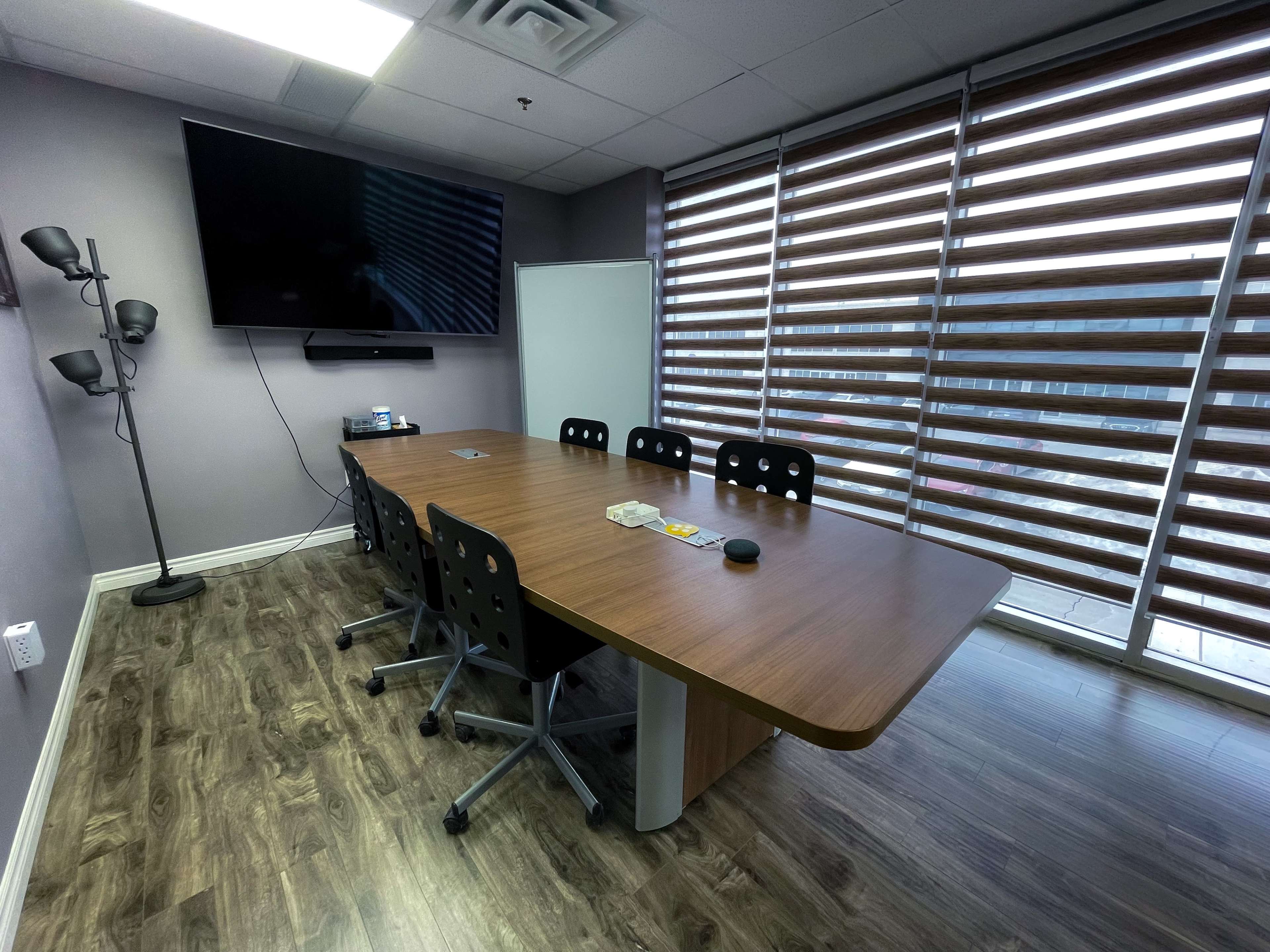 A conference room with a large wooden table, six black chairs, a television mounted on the wall, and window blinds partially open.