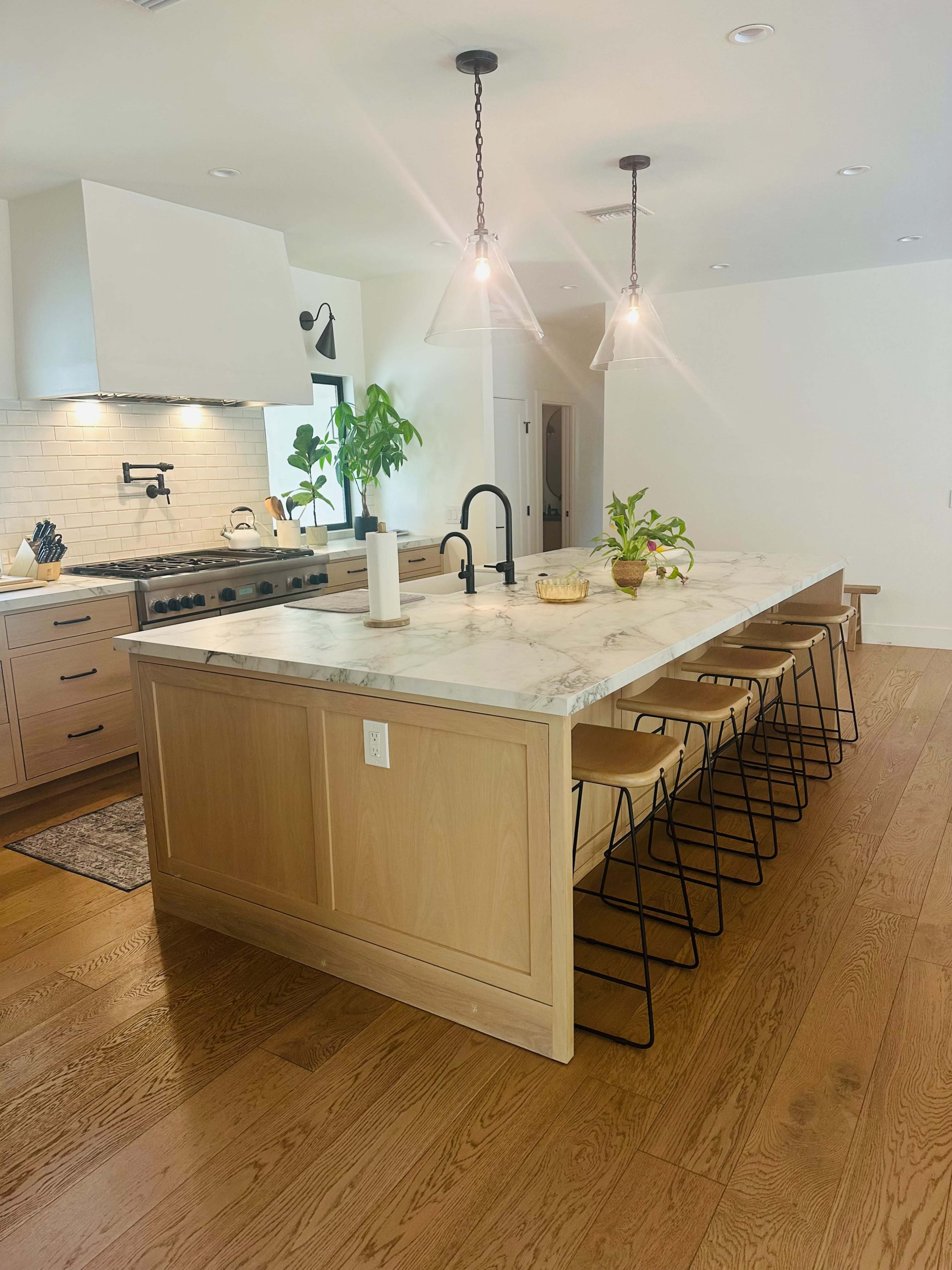 The image shows a modern kitchen featuring a large marble island with wooden cabinets, metal stools, and a white backsplash.