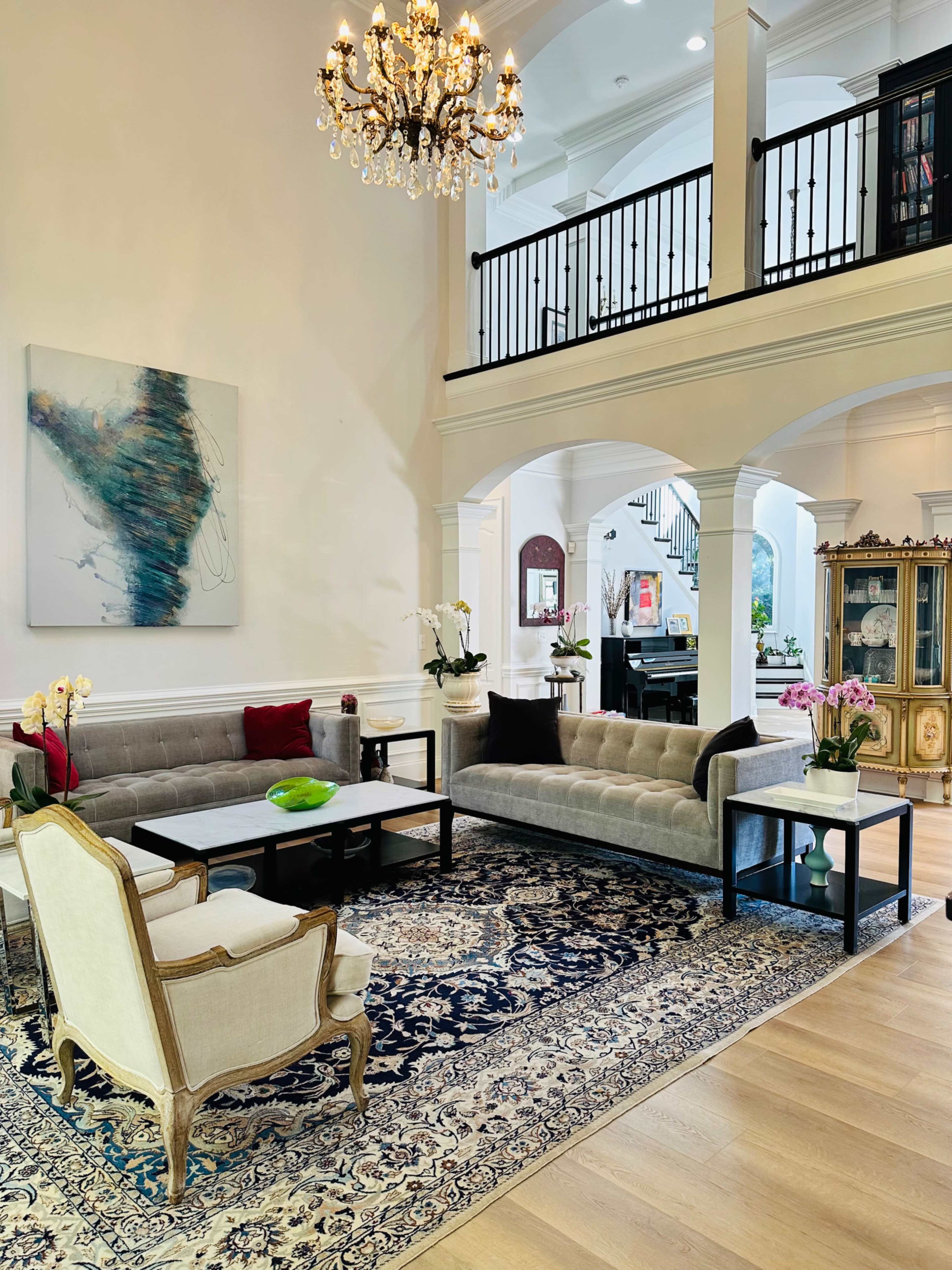 The image shows a spacious living room featuring a chandelier, a large artwork on the wall, two gray sofas, a marble coffee table, and a detailed rug.