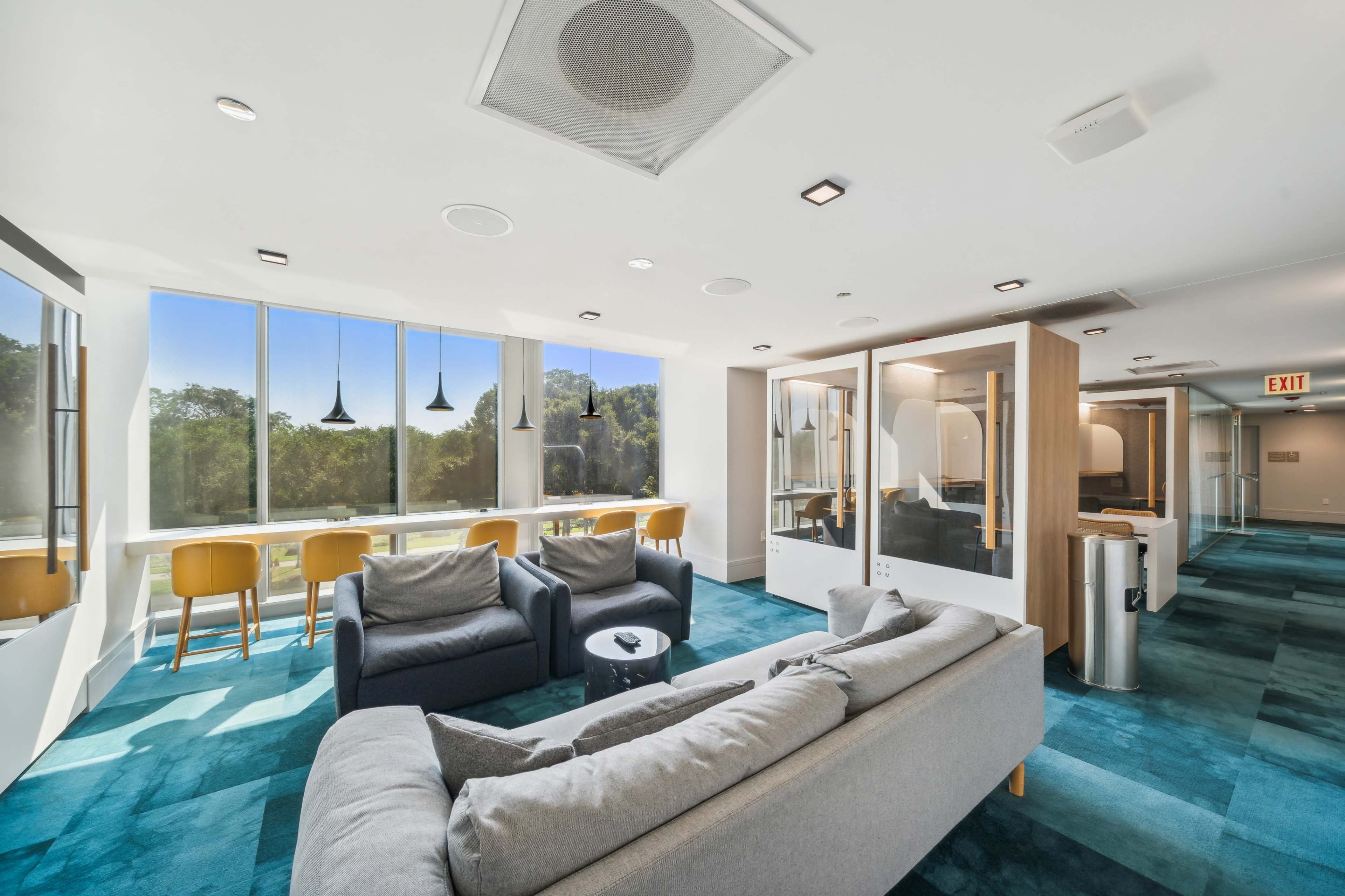 The image shows a modern lounge area with seating, large windows, and a view of greenery outside.