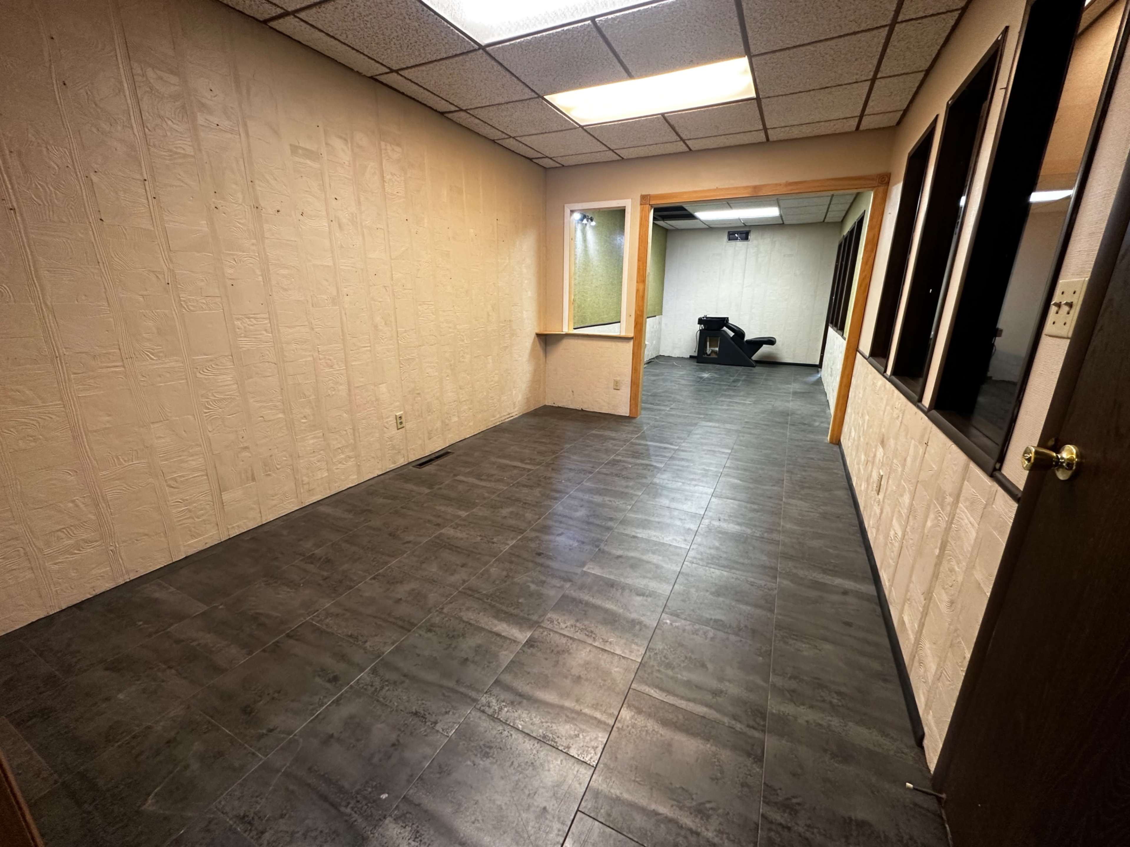The image shows a vacant room with textured walls, dark tile flooring, and a large window connecting to another area.