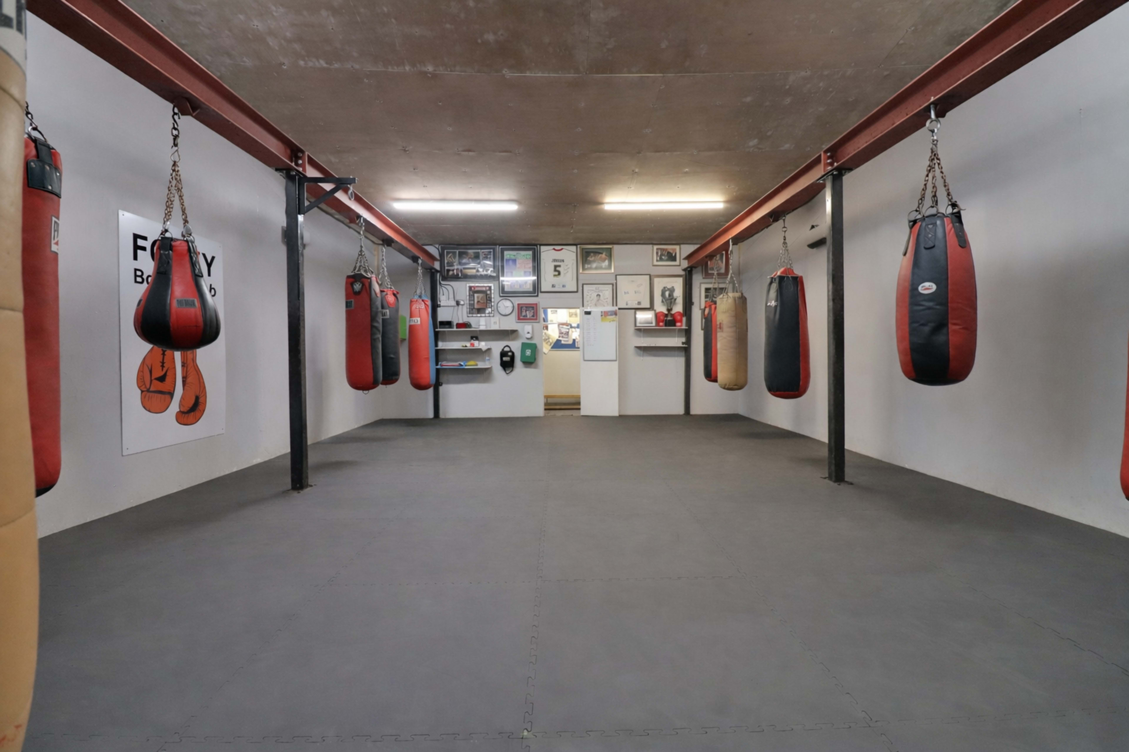The image shows an empty boxing gym with multiple hanging punching bags, black rubber flooring, and walls featuring various pictures and posters.