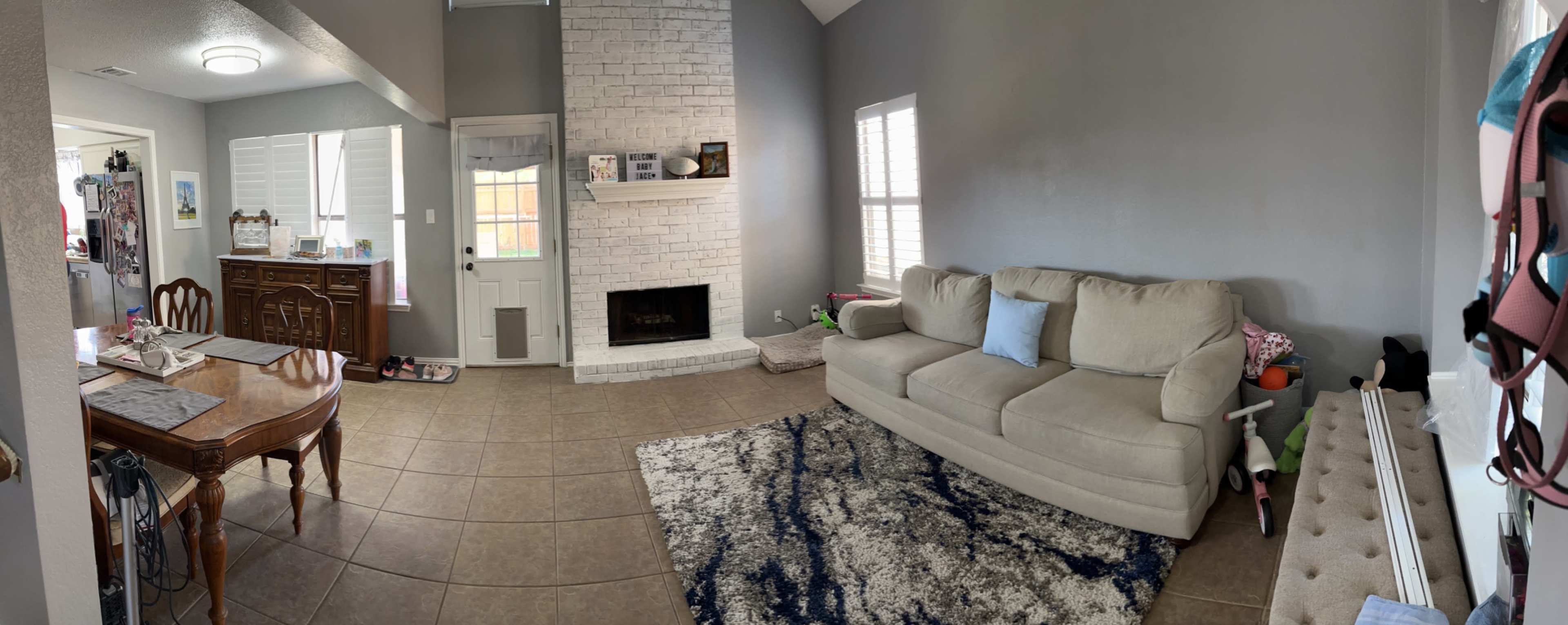 The image shows a living room with a gray wall, a beige sofa, a white brick fireplace, and a dining area with a wooden table.