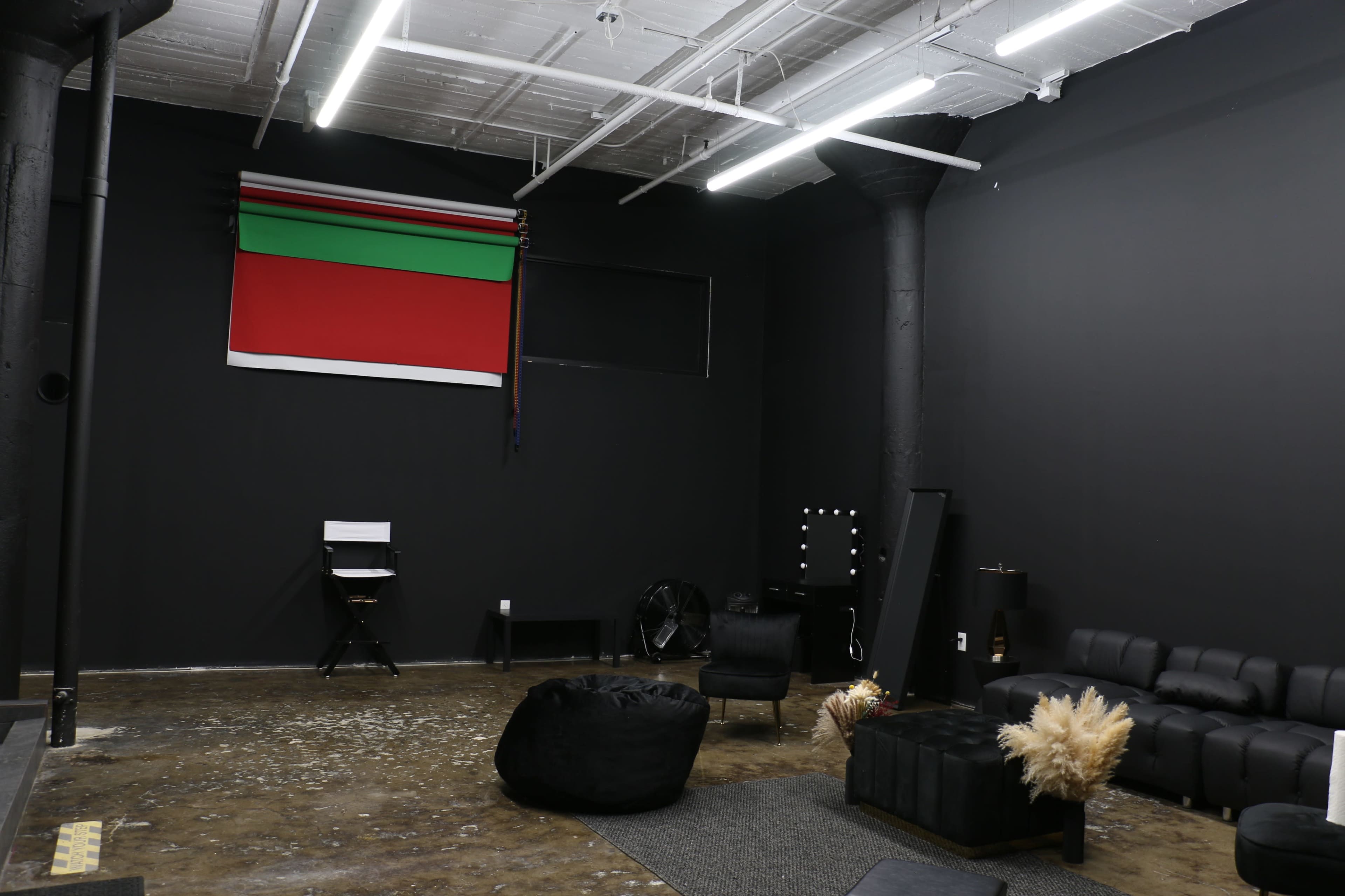 The image shows a spacious room with black walls, featuring minimal furniture, a large abstract flag on the wall, and a textured floor.