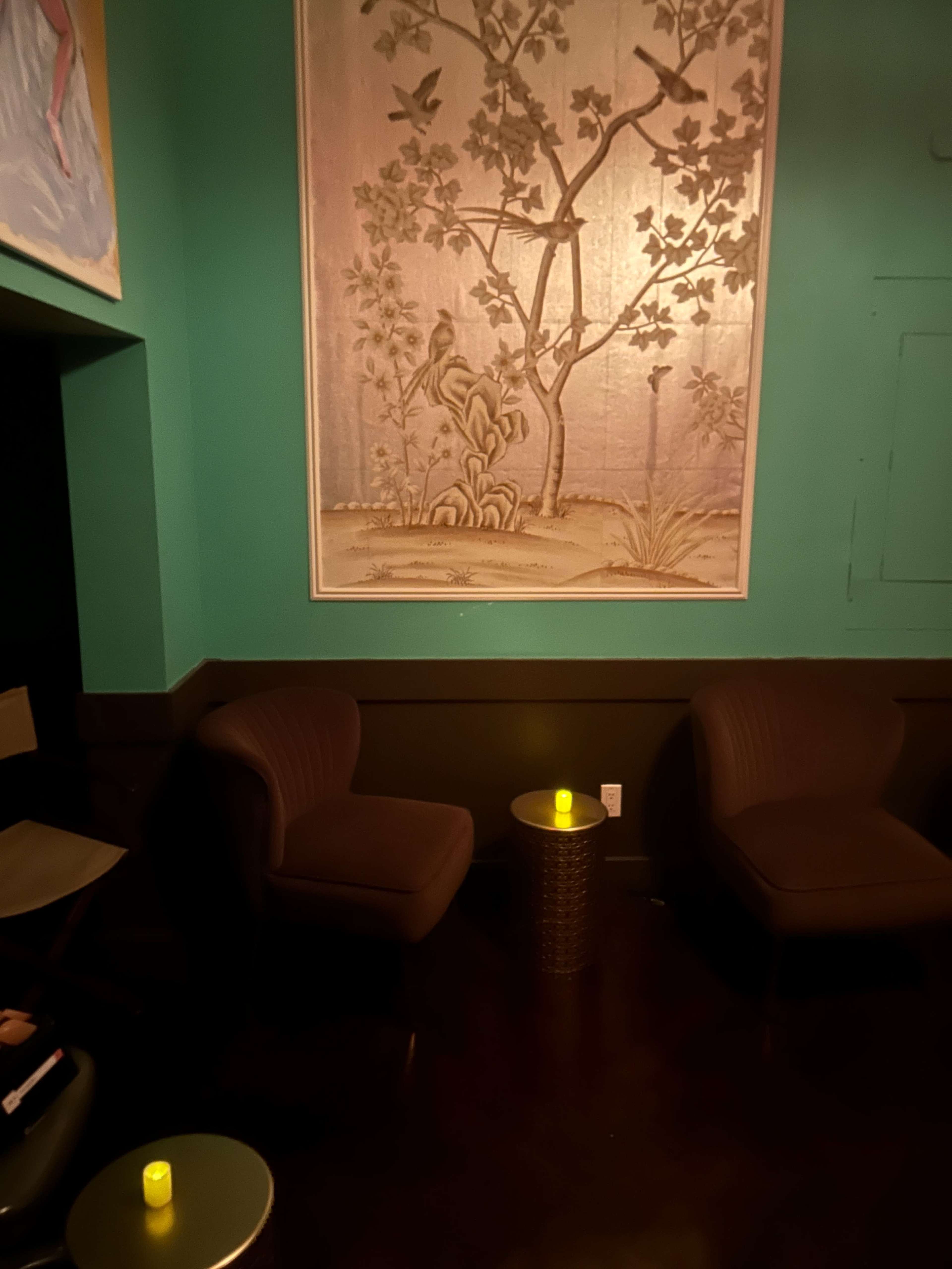 The image shows a dimly lit corner of a room with two dark chairs, a small table, and a decorative wall mural featuring trees and birds.