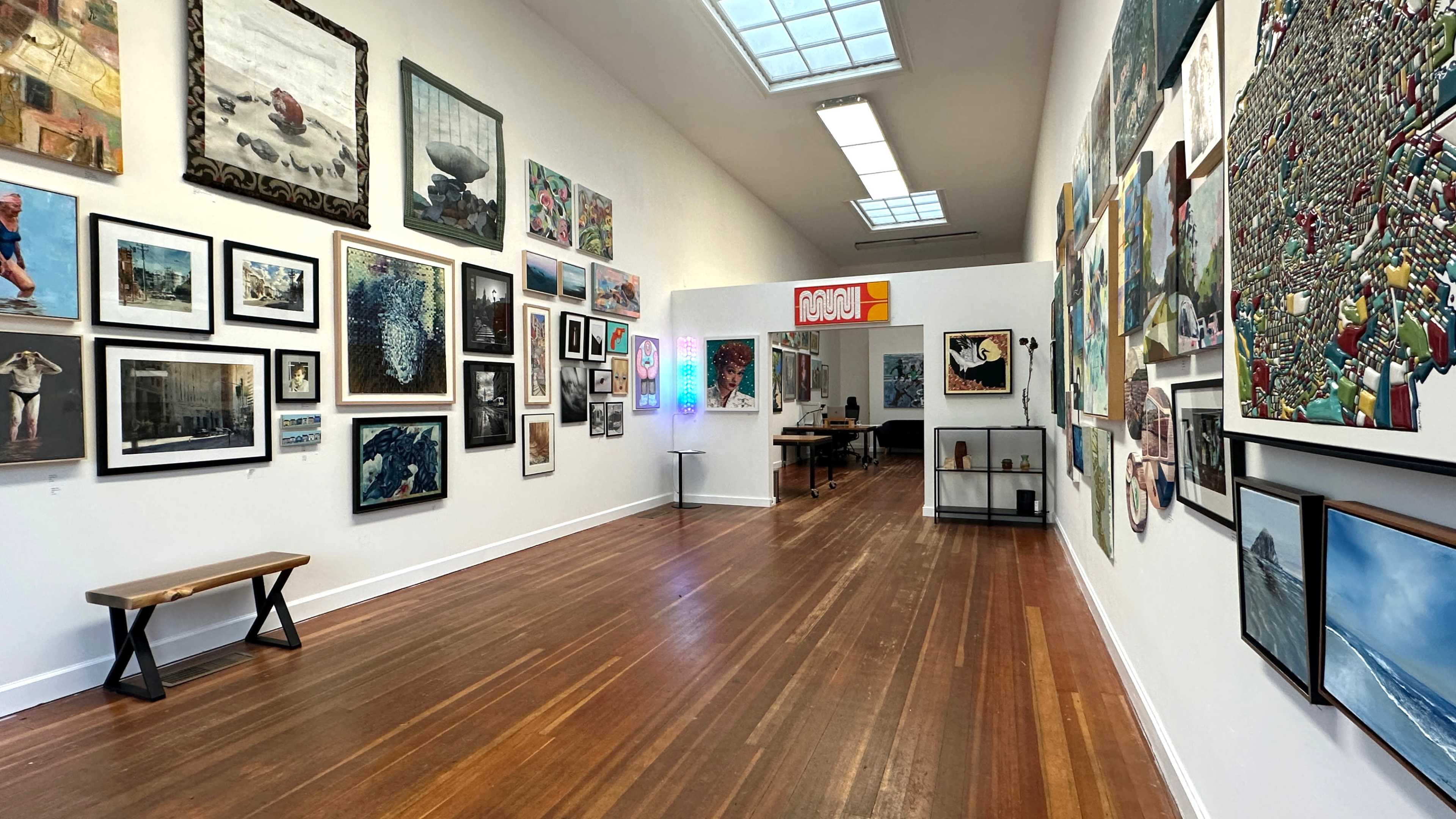 The image shows an art gallery interior with framed artworks displayed on the walls and a small seating area with a bench.