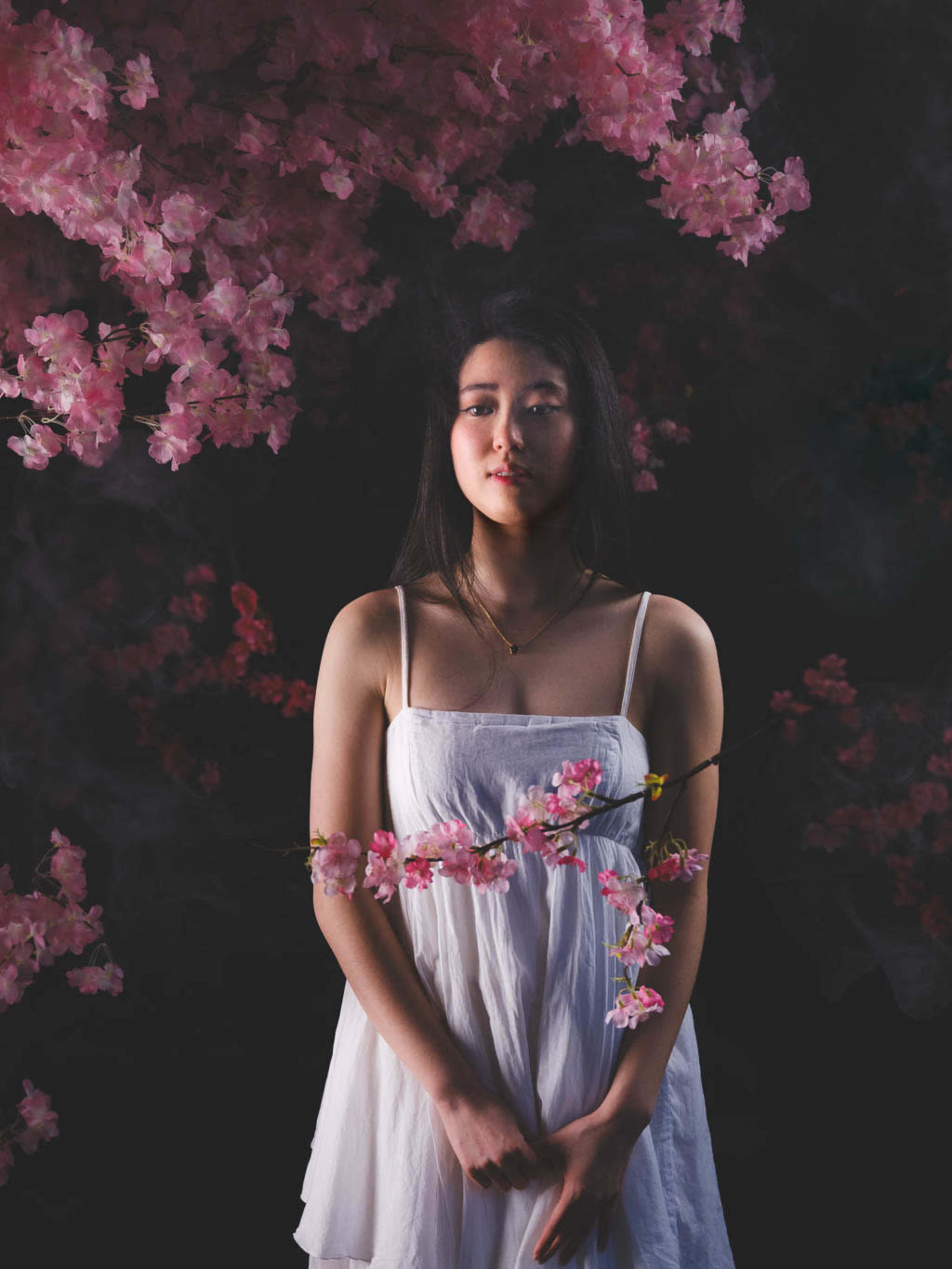 A woman in a white dress stands amidst cascading pink blossoms against a dark background.