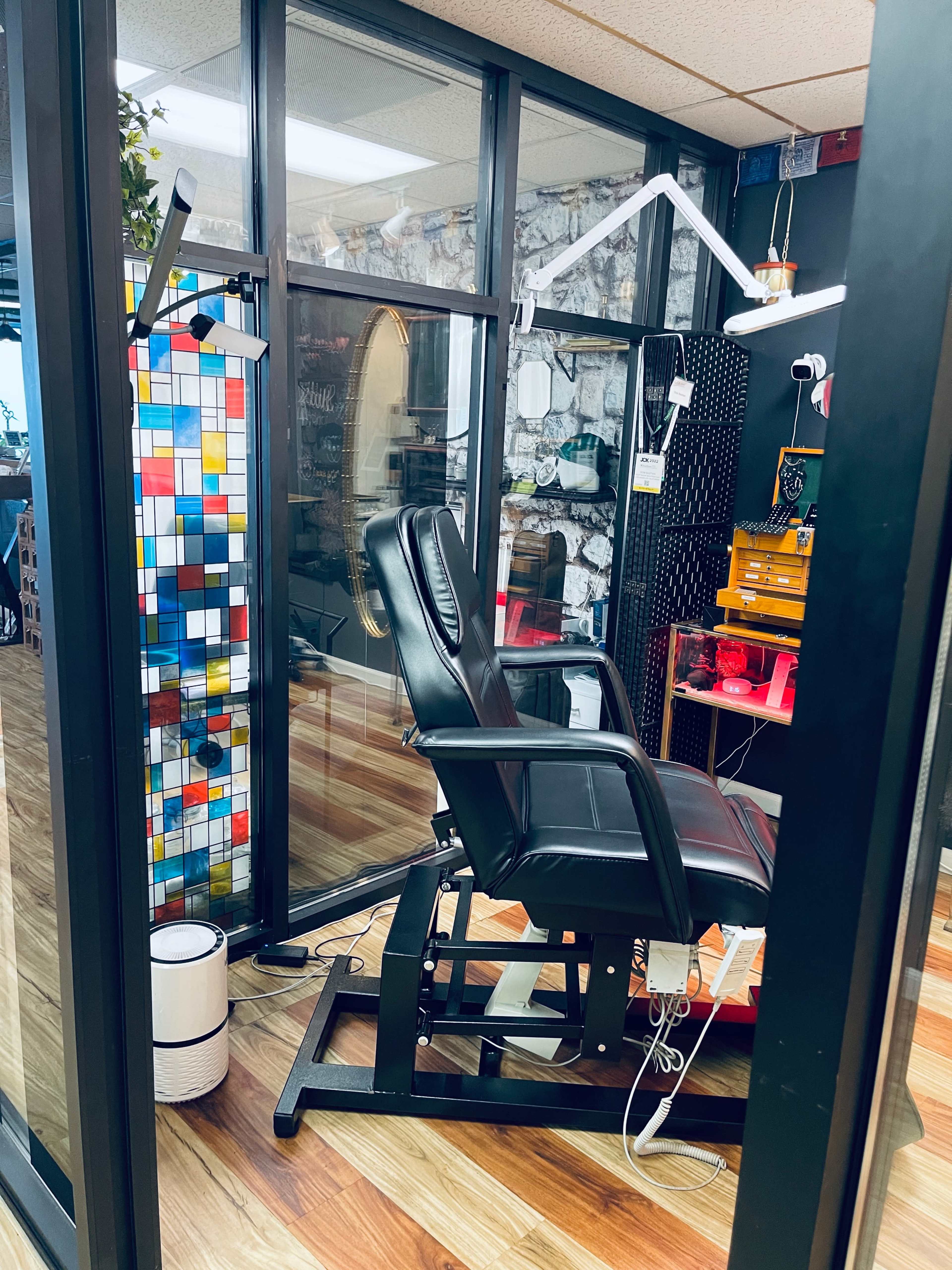 The image shows a tattoo studio with a black artist chair, colorful wall art in the background, and various tattooing tools and equipment displayed.