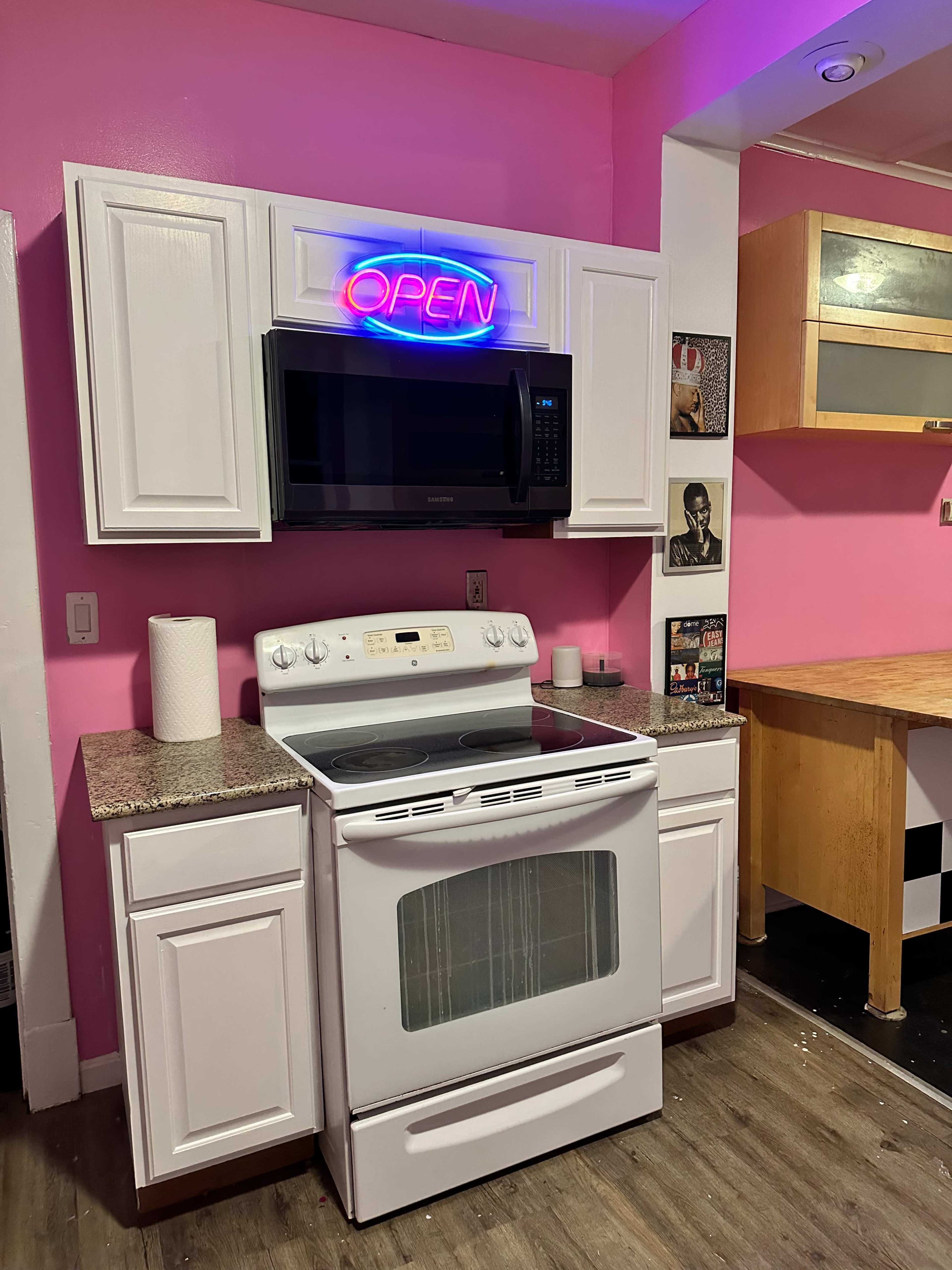 A kitchen features a white stove and microwave with a neon "OPEN" sign above, set against pink walls.