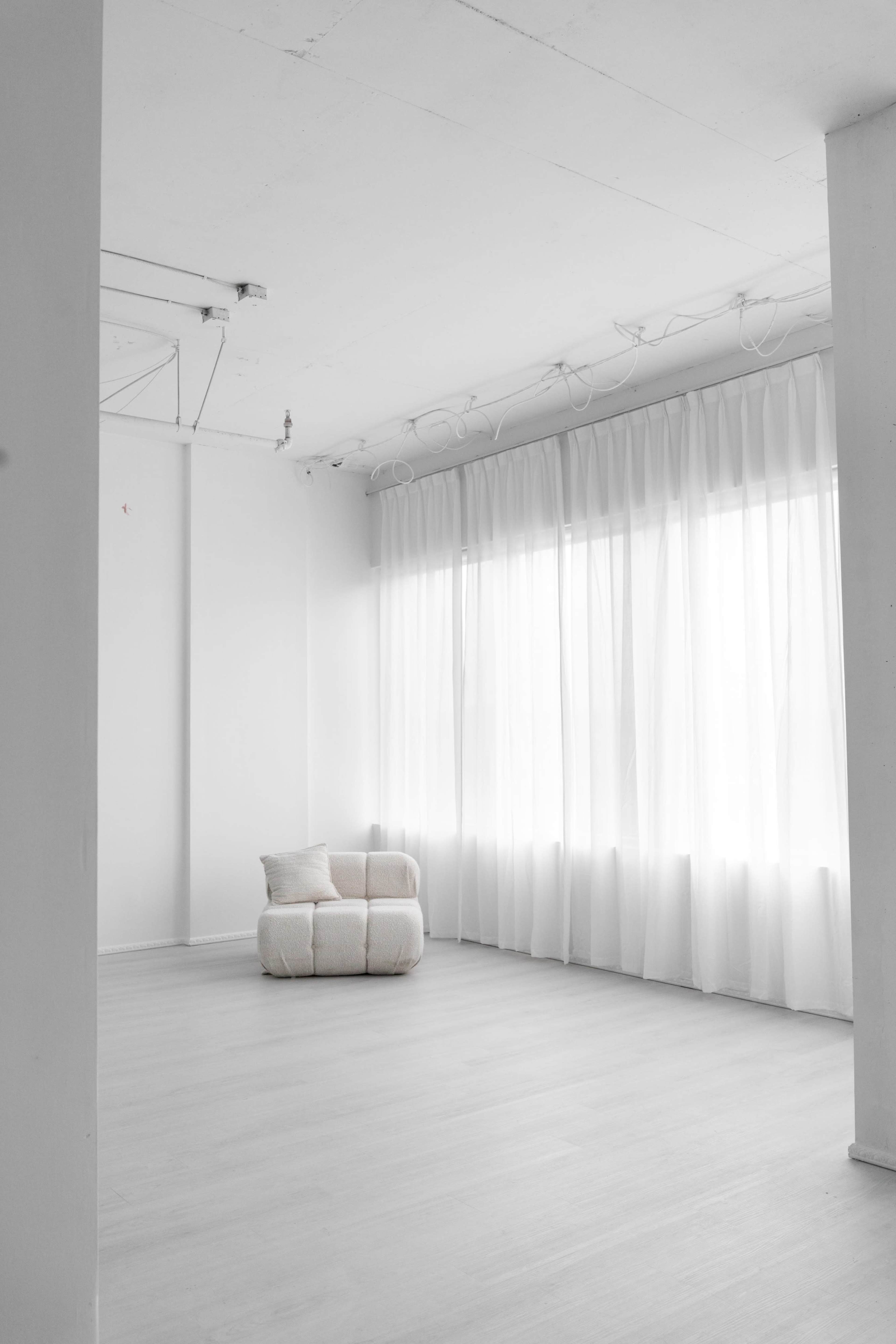 An empty room features a small, light-colored couch positioned near large windows covered with sheer curtains.
