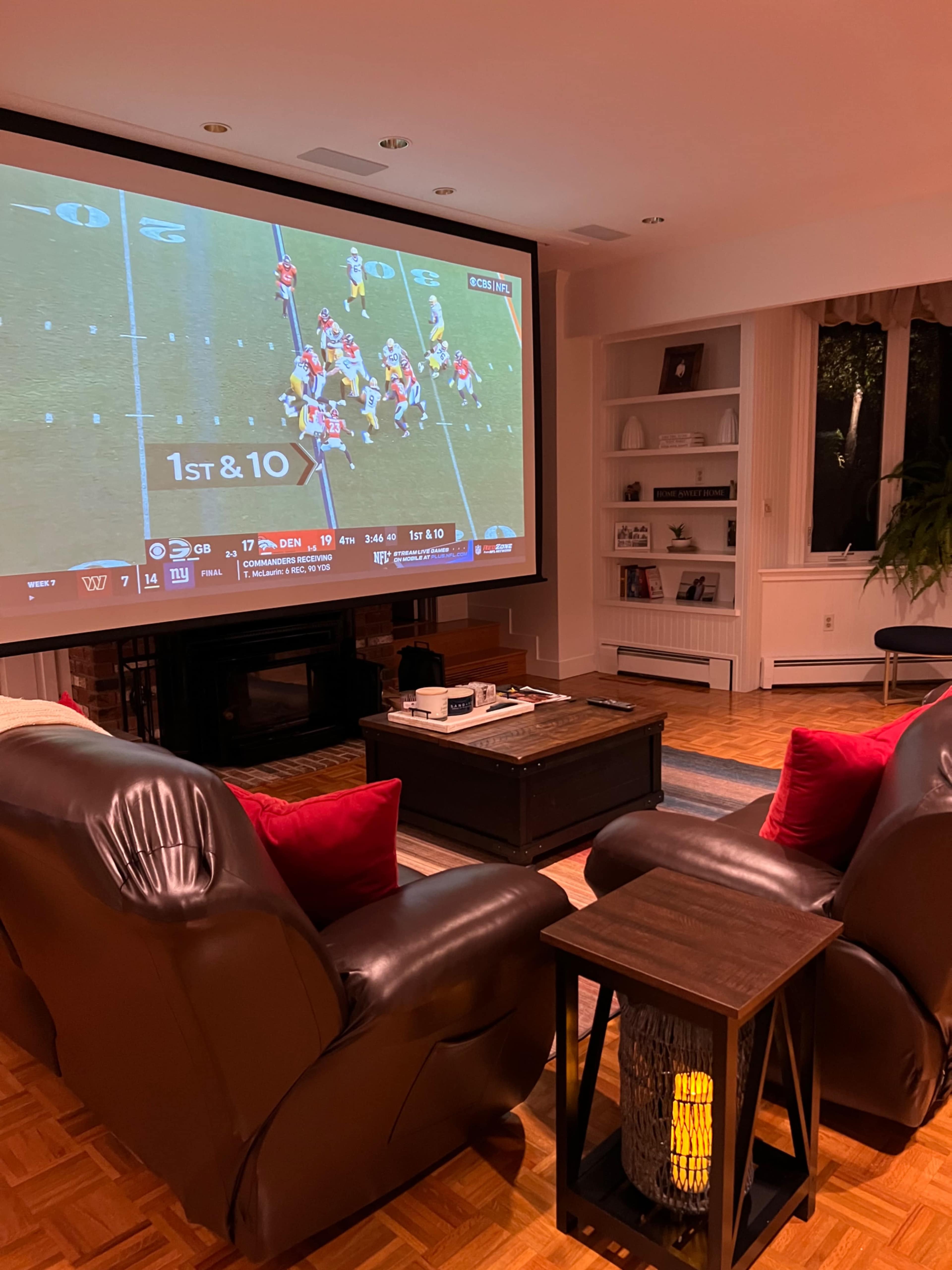 A cozy living room features two recliners facing a large screen displaying a football game.