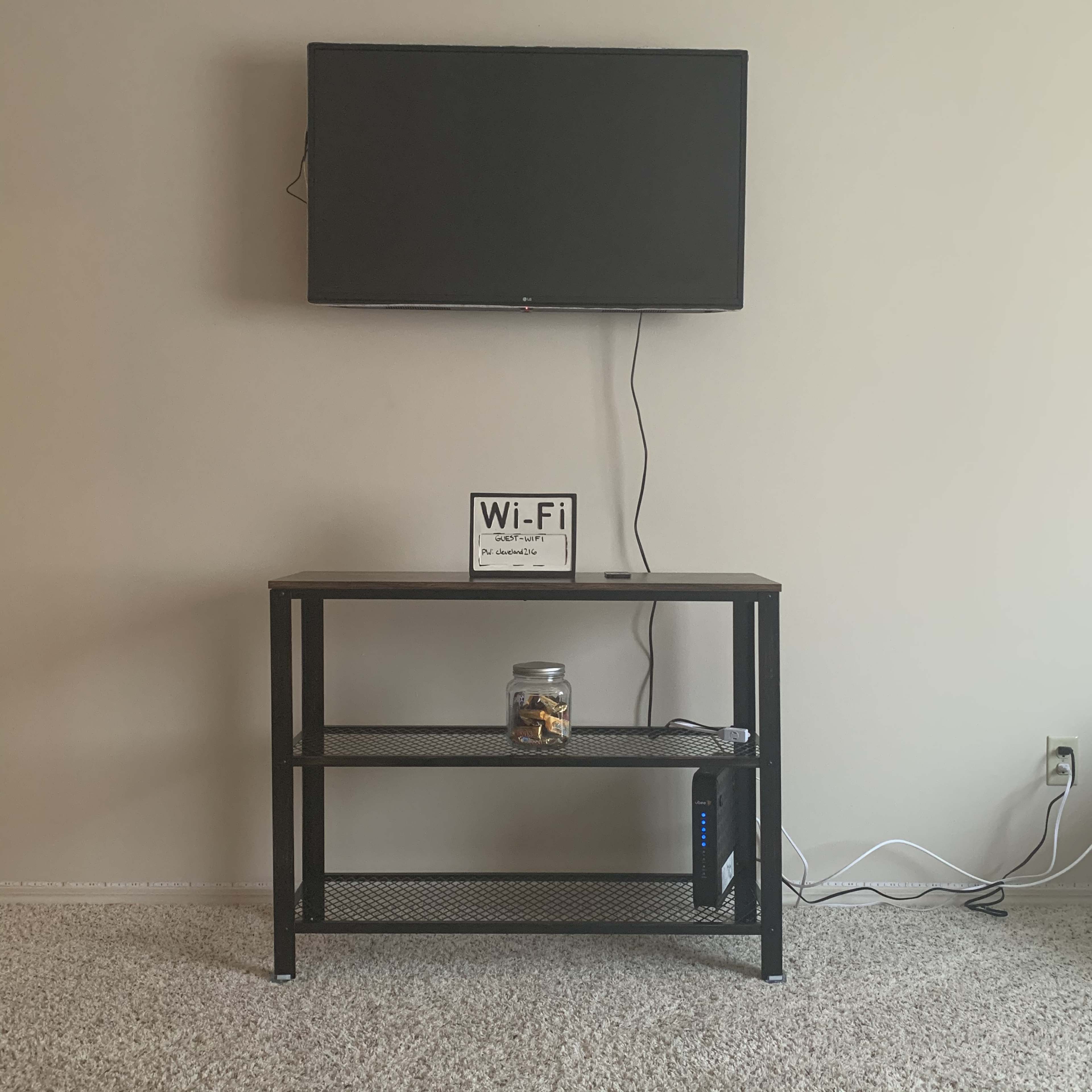 A flat-screen TV is mounted on a beige wall above a black metal stand, which holds a sign indicating Wi-Fi information and a jar, with a router placed underneath.