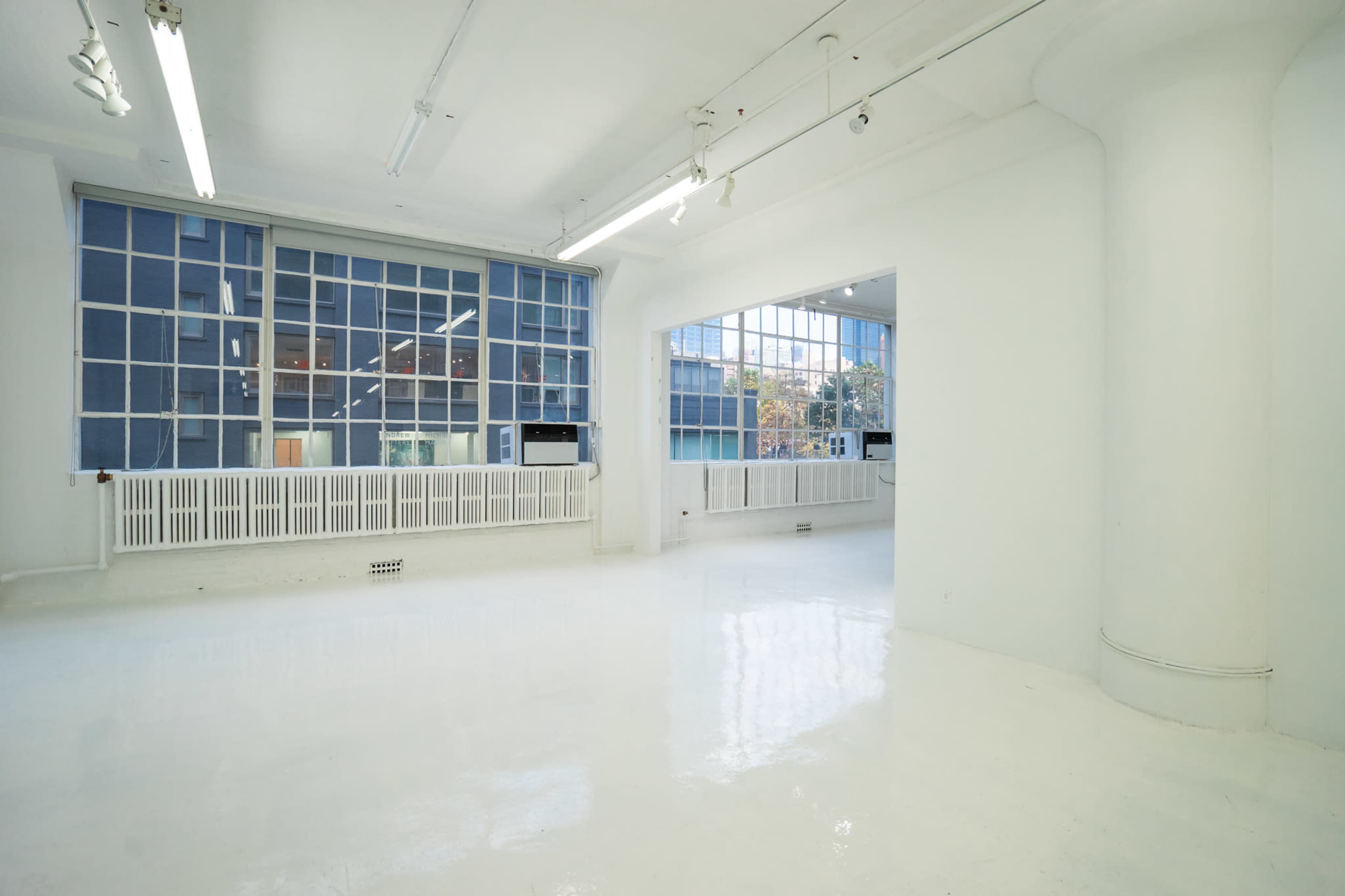 A spacious, empty room with white walls and flooring, featuring large windows that allow natural light to fill the space.