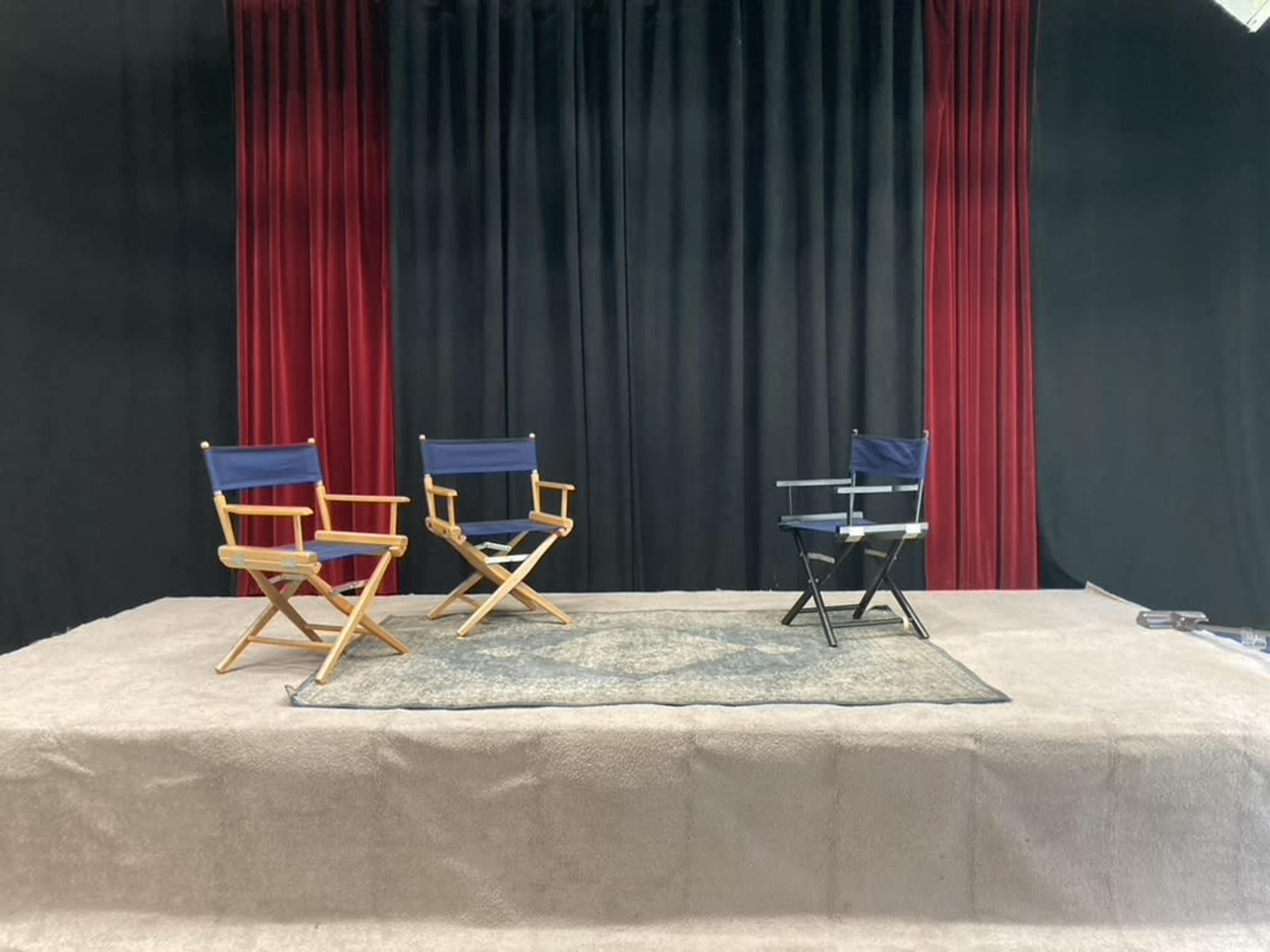 The image shows a stage with three director chairs on a rug, set against a backdrop of red curtains and black fabric.