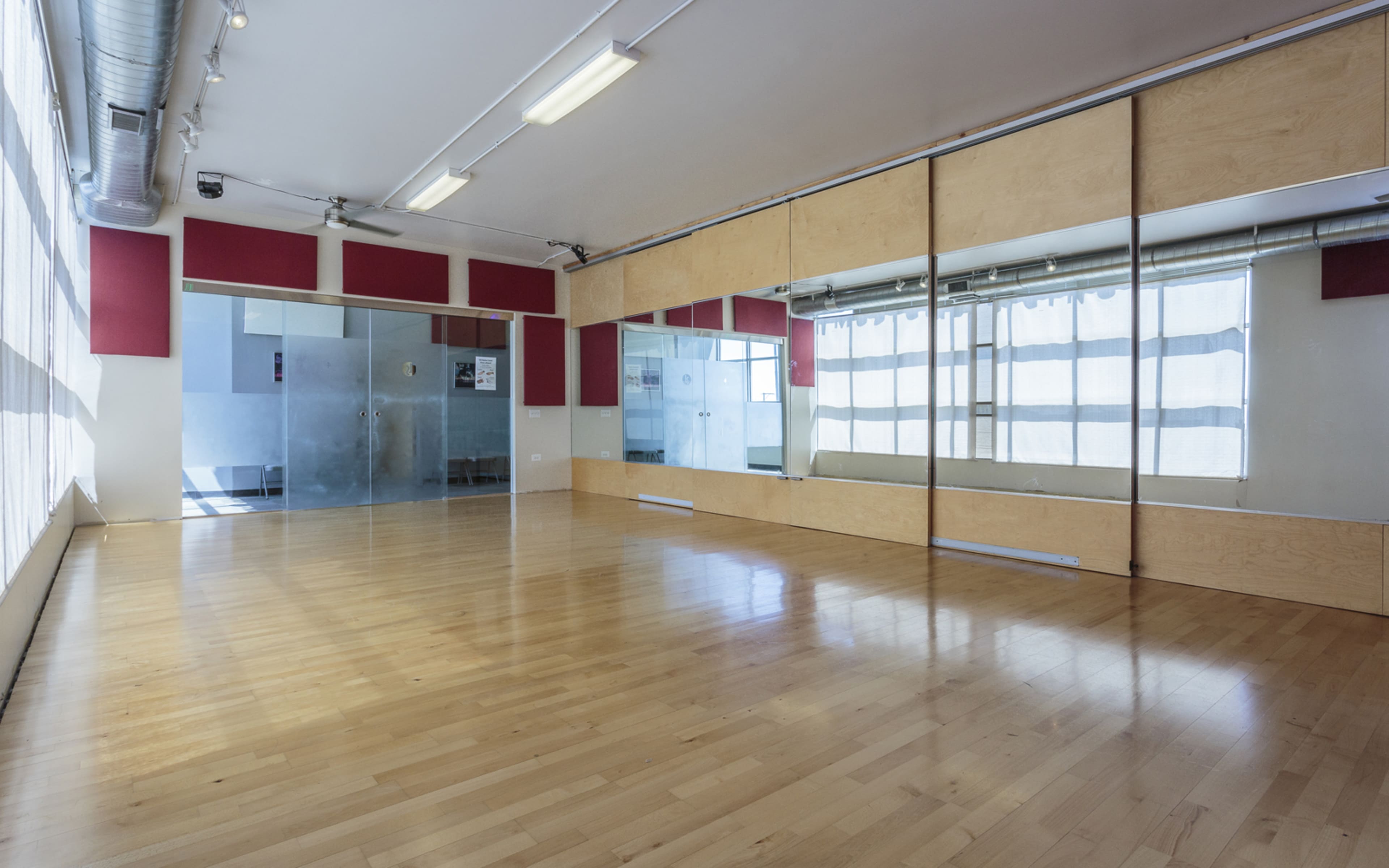 A spacious dance studio with wooden flooring, large mirrors on one wall, and natural light streaming through windows covered with sheer curtains.