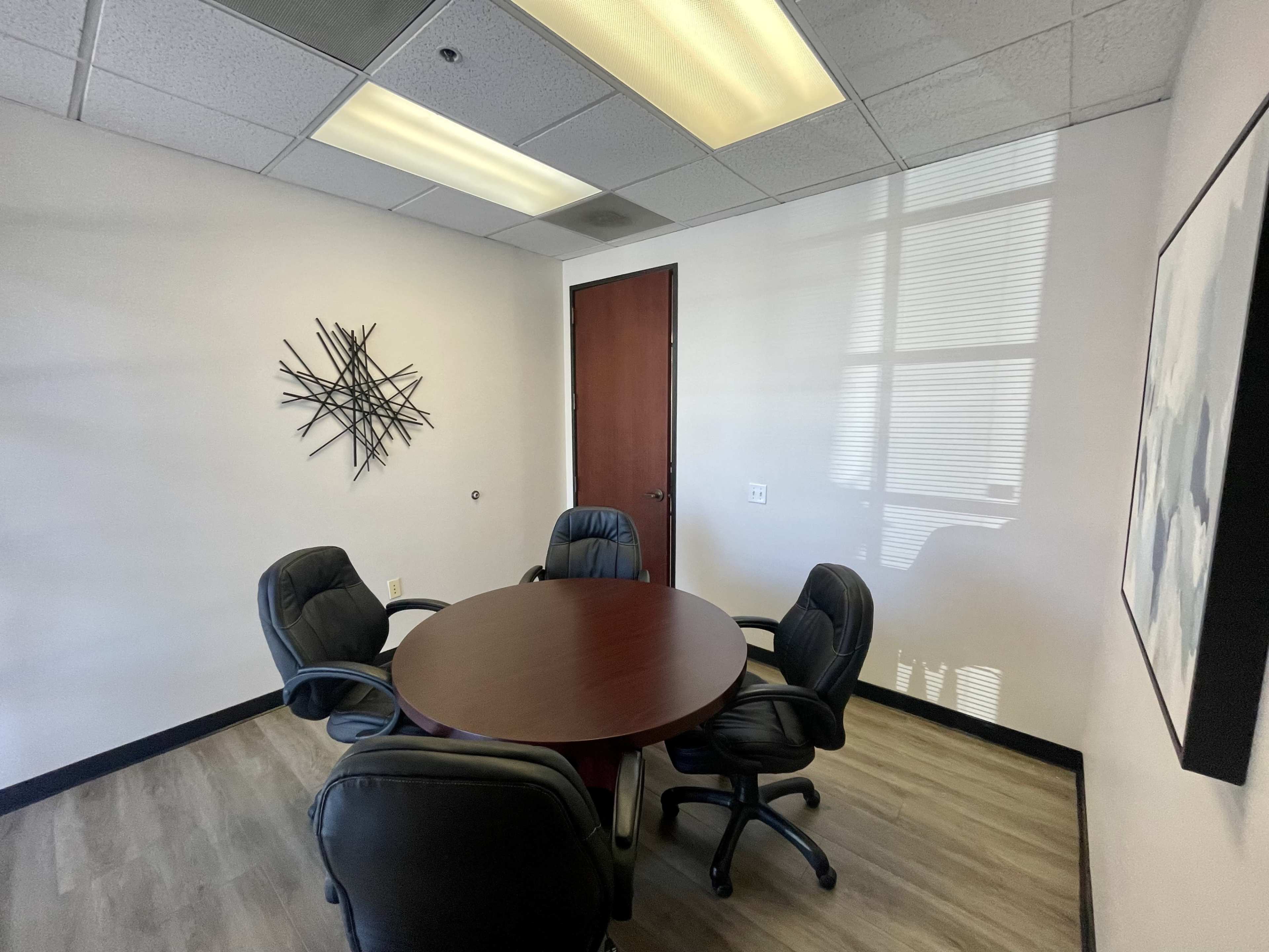 A small conference room features a round table surrounded by four black leather chairs, with a wall art piece and a wooden door.