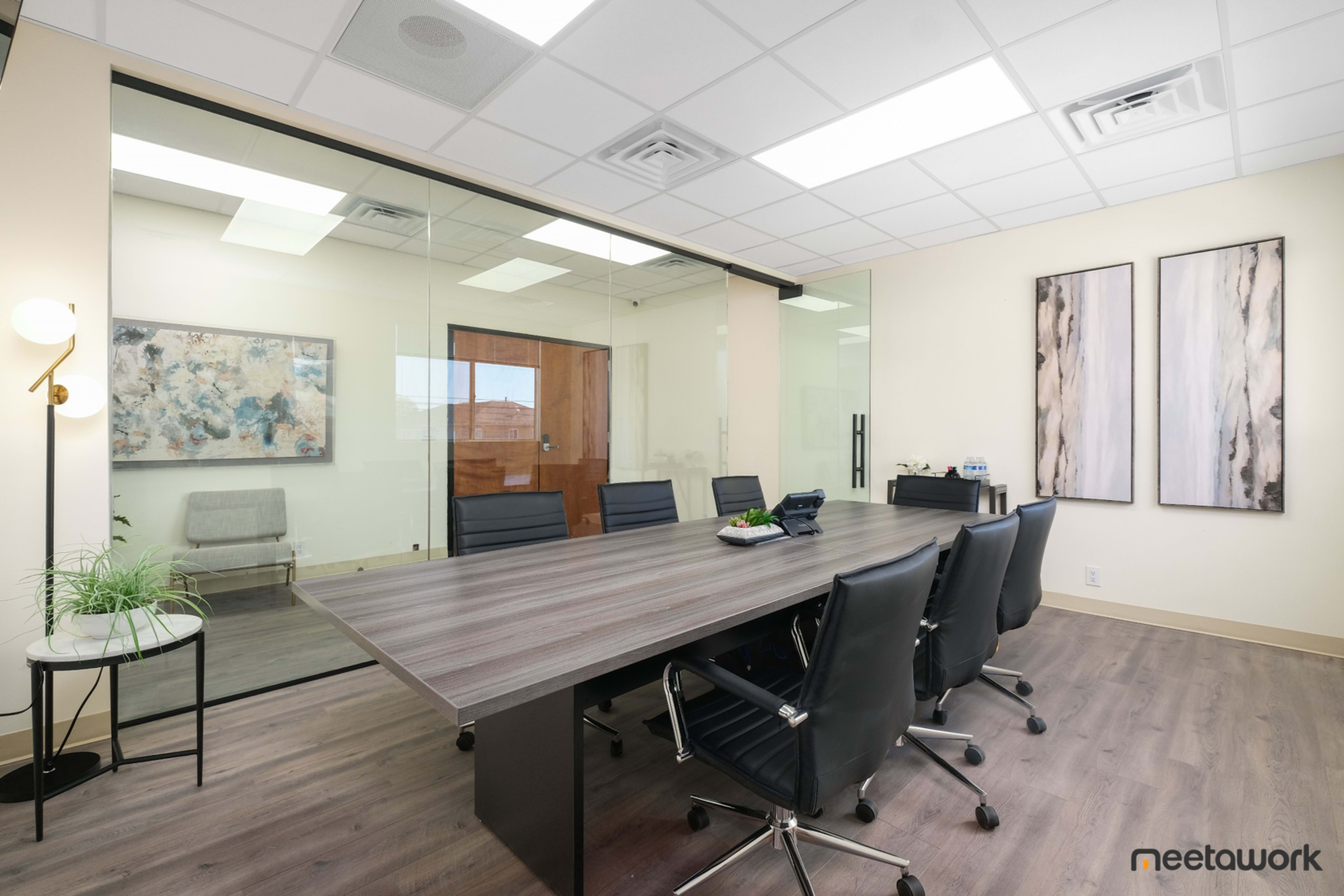 A modern conference room features a large wooden table surrounded by black chairs, with glass walls and artwork on the nearby walls.