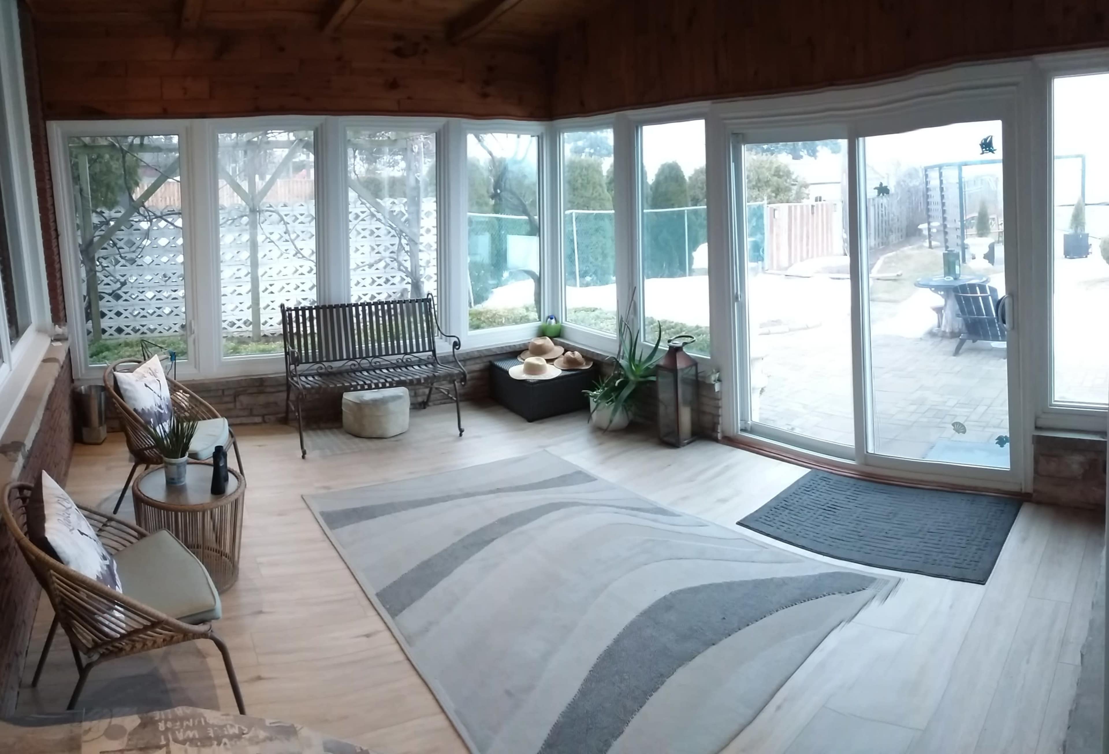 A sunroom with large windows, a wooden ceiling, and seating arrangements including a bench and chairs, alongside a rug on the floor.