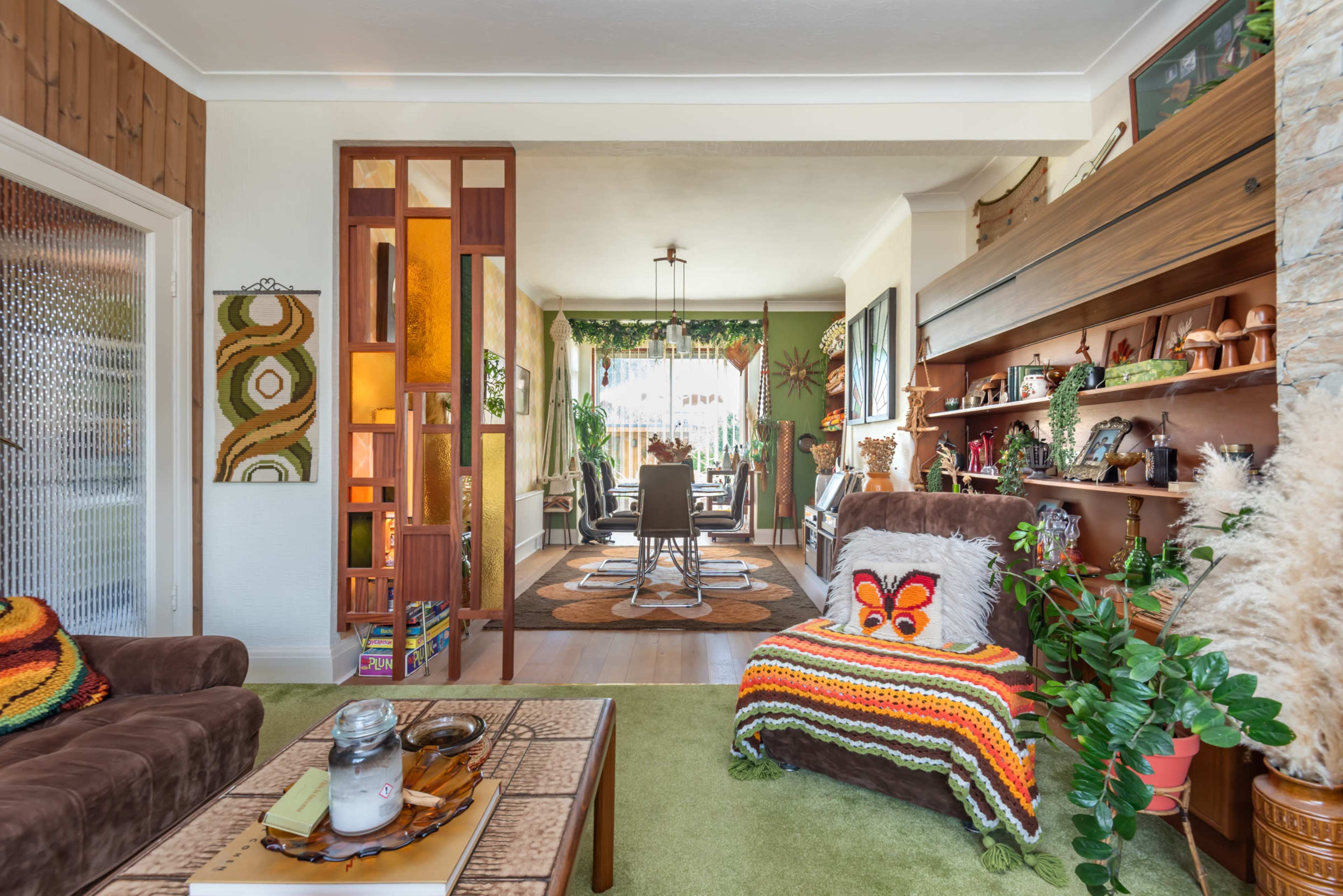 The image shows a living room with a brown sofa, a colorful crocheted blanket, various plants, and a partition leading to a dining area filled with natural light.