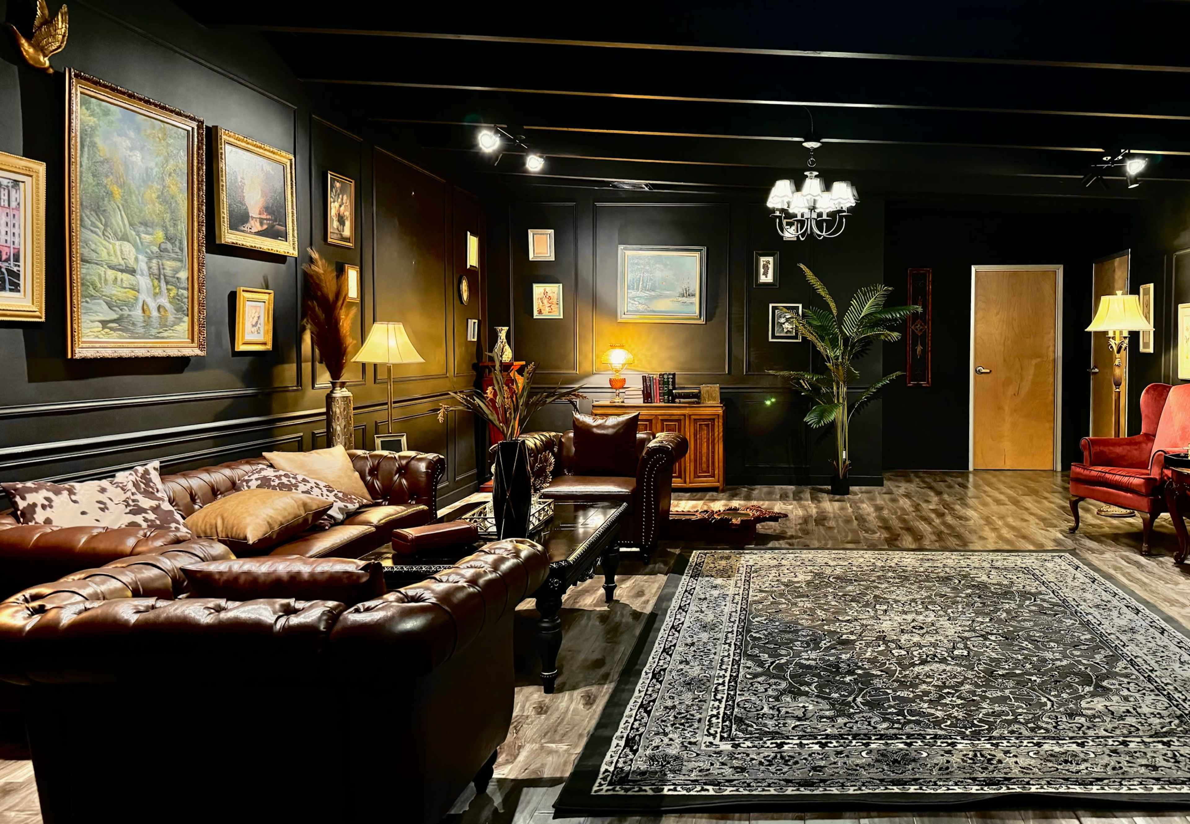 The image shows a dimly lit living room with dark walls, a leather sofa, a wooden cabinet, framed paintings, and a decorative rug.