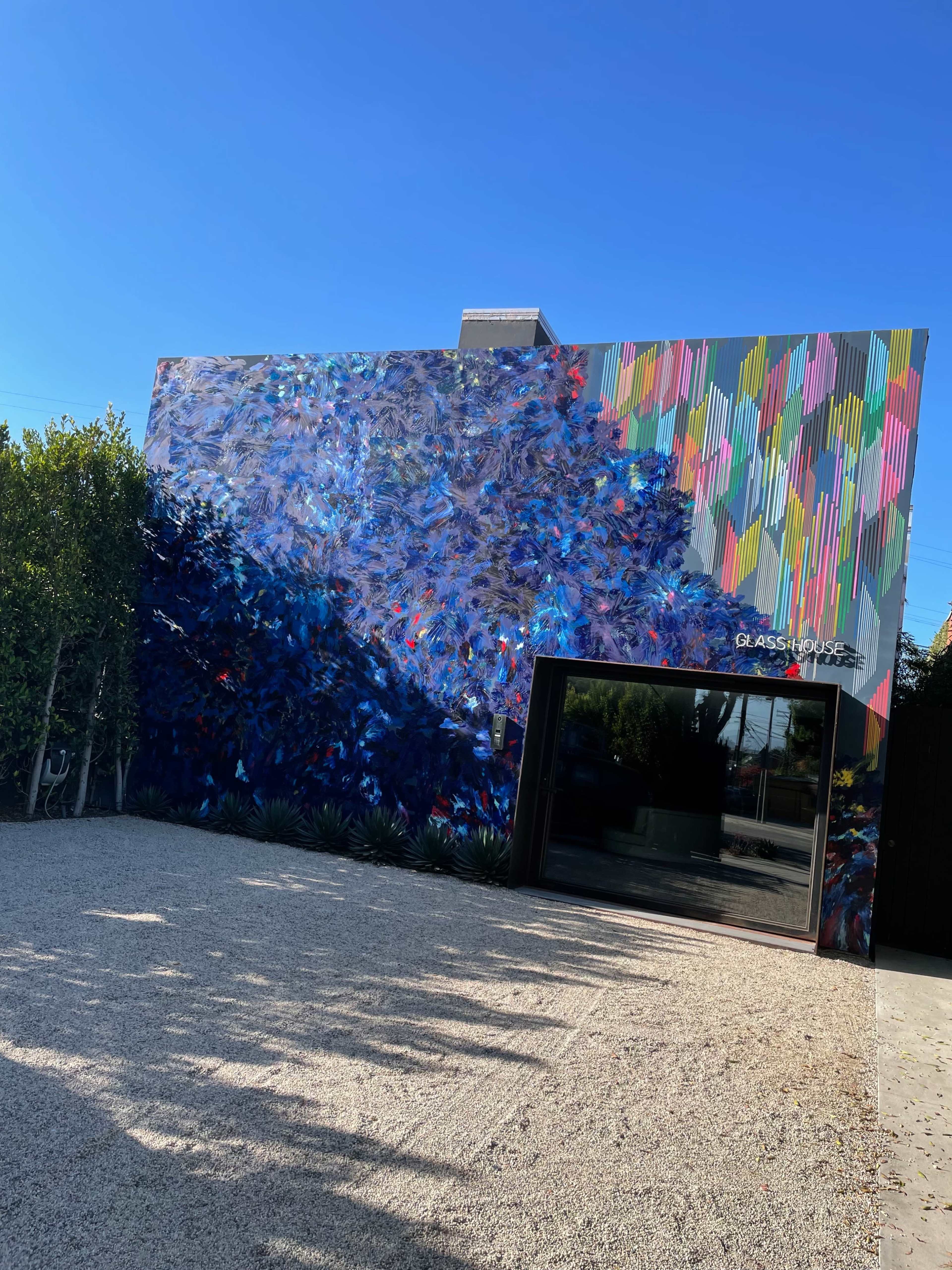 The image shows a brightly painted mural on a building facade with a modern entrance and a gravel driveway.