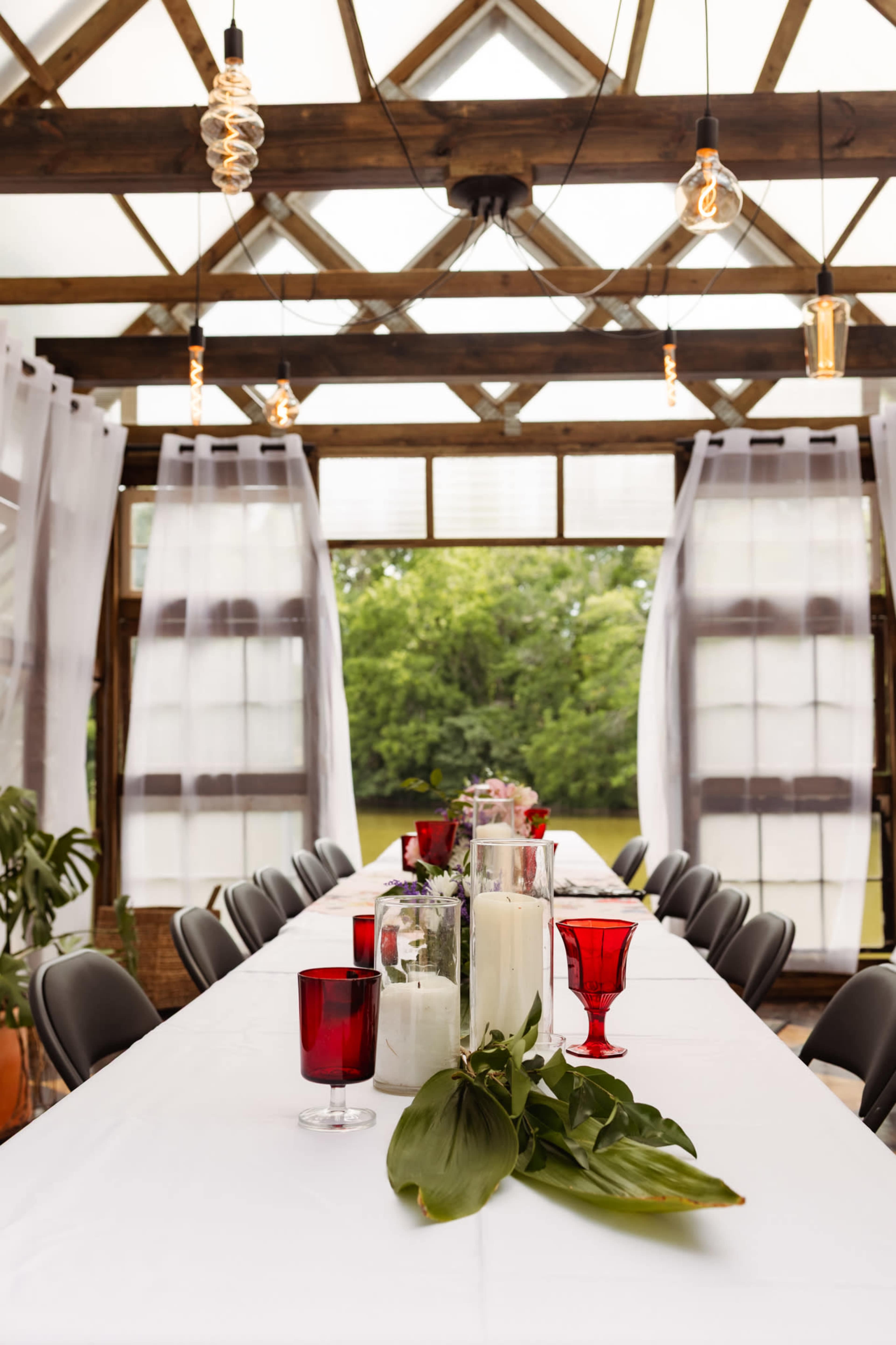 A long dining table is set with red and clear glass vases, surrounded by chairs, and features large windows that open to a greenery view.