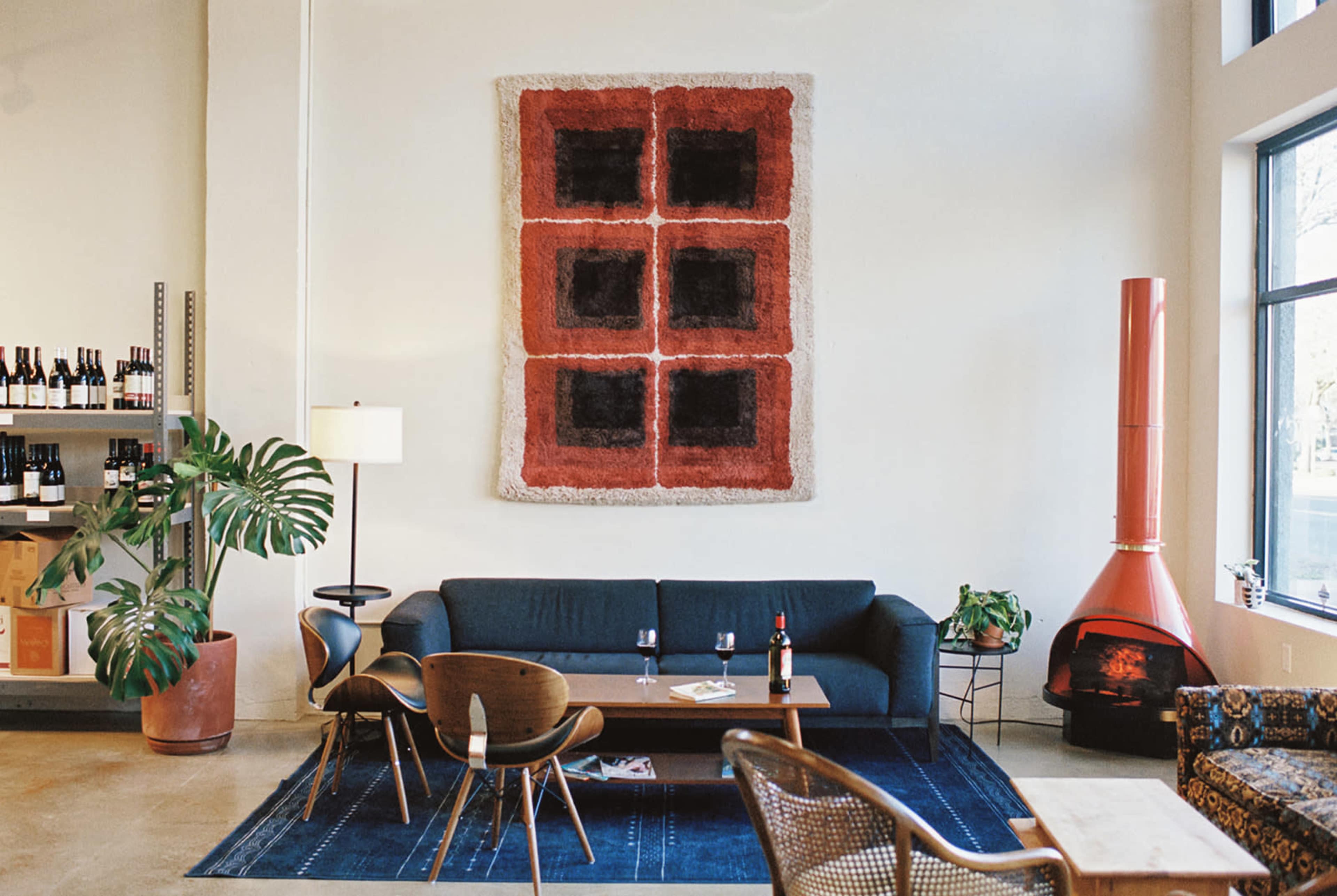A modern interior space features a large textile artwork on the wall, a dark sofa, a wooden coffee table, and a red fireplace, with bottles of wine displayed on shelves in the background.