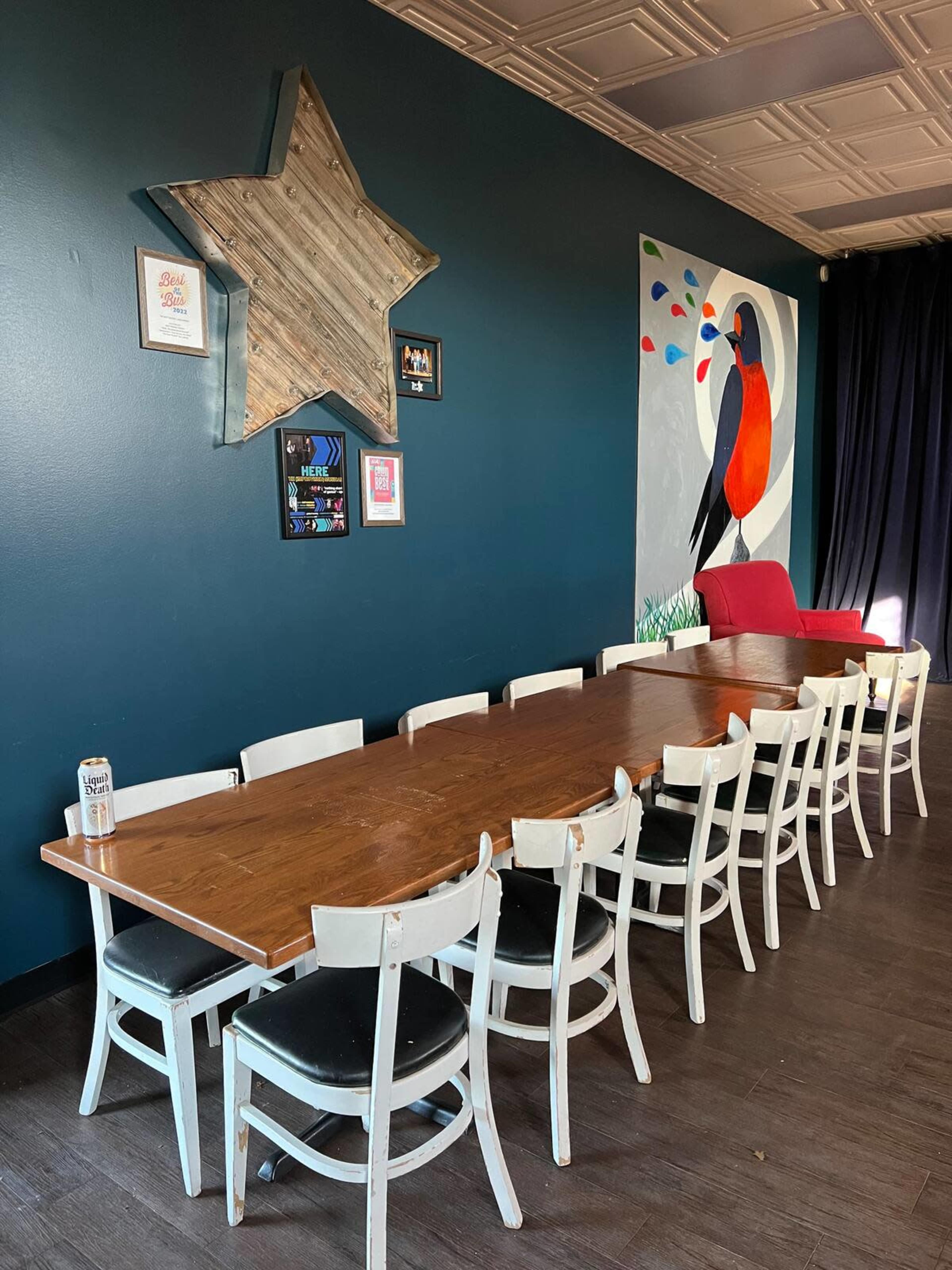 A long wooden dining table is flanked by white chairs in a room featuring a dark blue wall with a star decoration and a colorful bird mural, along with a red chair in the background.
