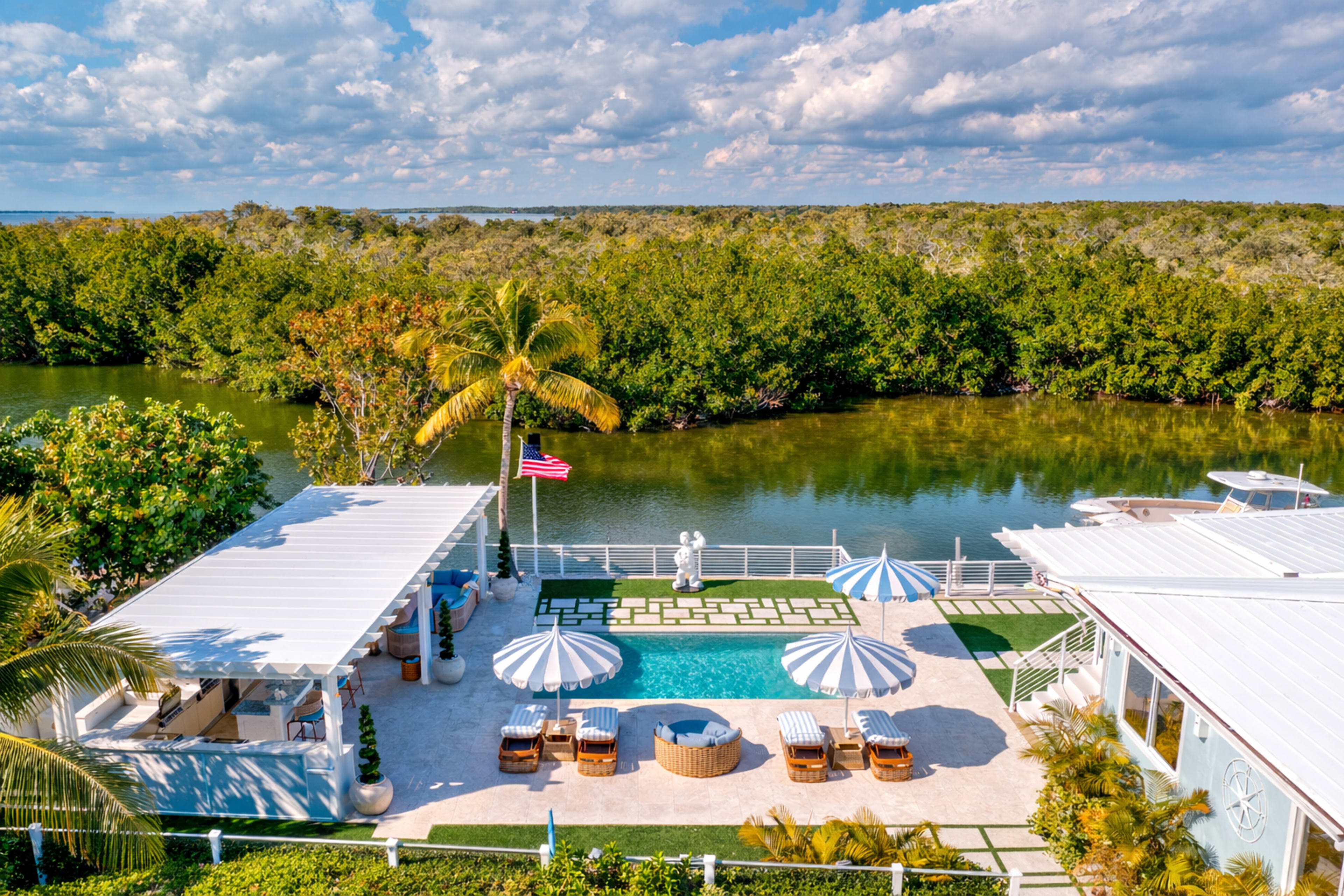 The image shows a luxurious waterfront property with a swimming pool, outdoor seating area, and palm trees, surrounded by lush greenery and water.